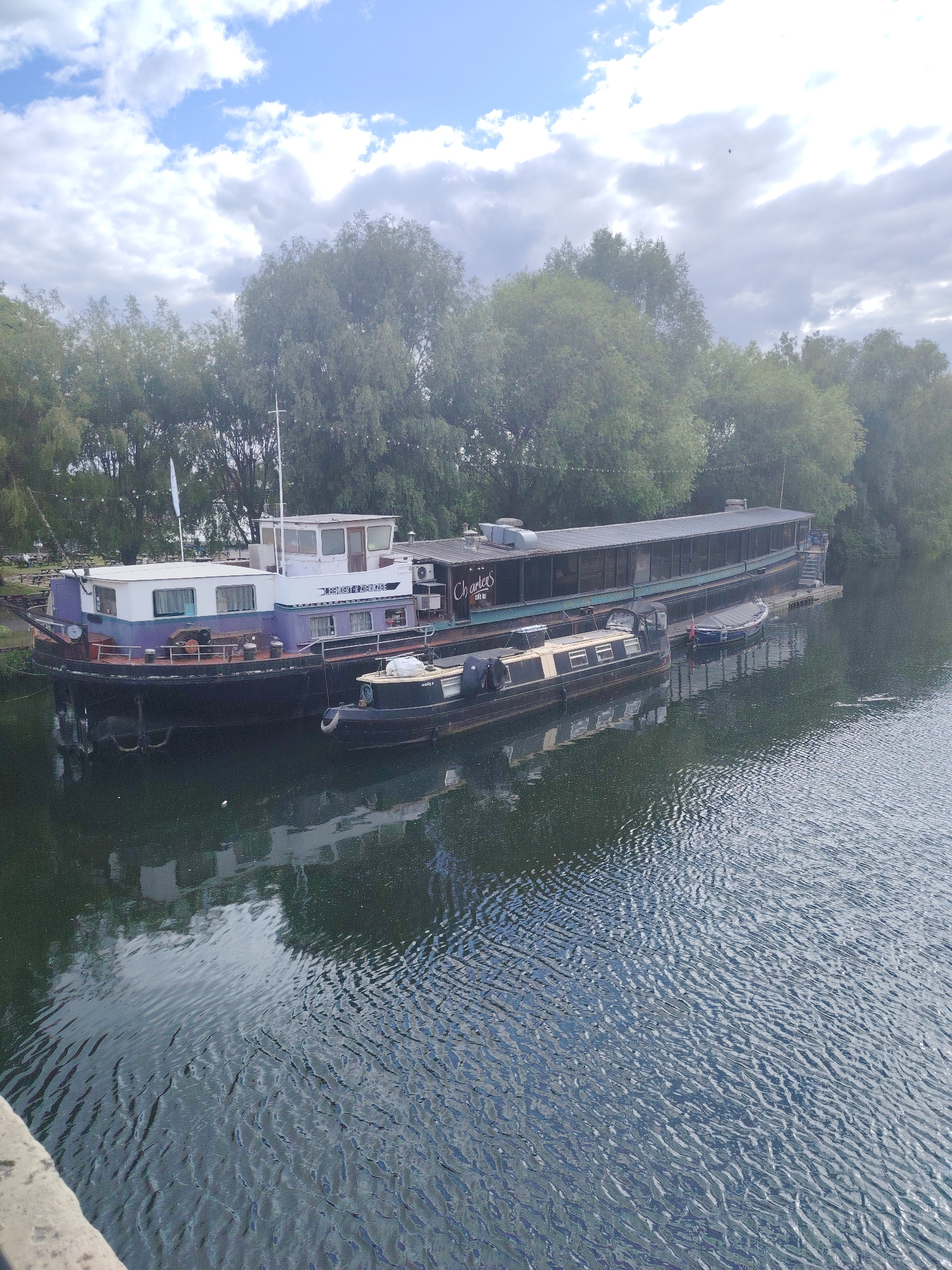 The image depicts a daytime scene in Peterborough, United Kingdom, showcasing several moored vessels on a body of water, likely a river or canal. In the foreground, the edge of a concrete structure, possibly a bridge railing, is visible on the lower left. The water surface exhibits numerous ripples and reflects the sky and adjacent structures.
Centered in the image is a large, two-story boat painted purple and white with a black hull. Its superstructure is white, and the hull below the waterline is black. The vessel features a pilothouse, multiple windows, and a mast with a white flag. Text on its side reads "EBENDERY ZIERZEE". To the right of this boat, and extending further along the waterway, is a very long, dark-hulled barge-like vessel. This vessel appears to be converted, featuring a prolonged enclosed upper deck with numerous windows along its length. A partial sign on its side is visible, reading "Charters BAR BUT". Moored alongside this longer vessel are two smaller boats: one dark-hulled narrowboat with cream-colored detailing and another smaller dark vessel further down the bank.
The far bank of the waterway is densely lined with mature green trees. Beneath the trees, behind the purple and white boat, several picnic tables are discernible, suggesting a recreational area. The sky above is partially cloudy with patches of blue visible. A small, dark bird is captured in flight in the upper central portion of the sky.