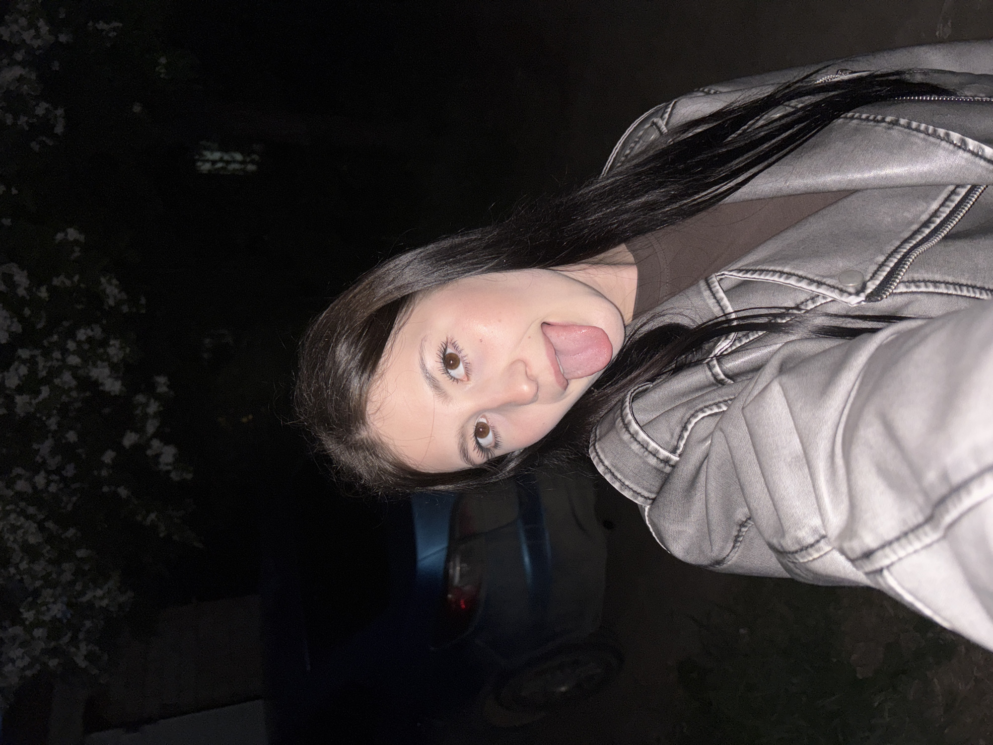 A young woman is seen in a selfie-style image, sticking her tongue out. She has dark hair and brown eyes, and is wearing a gray jacket. The background is dark, suggesting it is nighttime, with some faint greenery visible on the left and a dark-colored car partially visible behind her. There's insufficient information in the image to determine the precise location within Фоки, Russia.