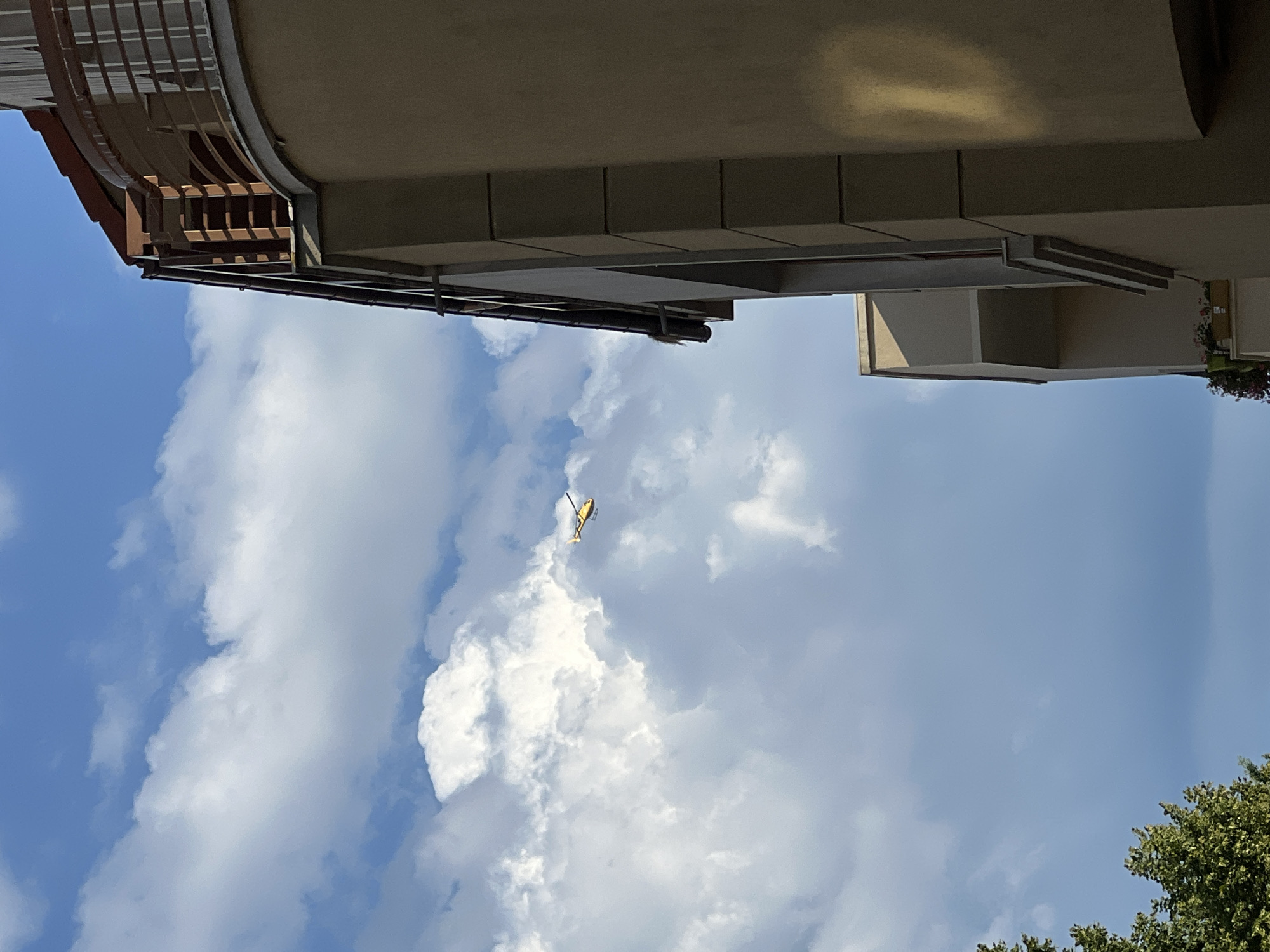 A bright yellow toy helicopter is visible in the sky, flying near white clouds. The helicopter is small and appears to be a drone. The sky is blue and partly cloudy. A building with tan walls and a brown roof is partially visible on the right side of the image. The building has a balcony with a railing. The scene appears to be set in a city, possibly Constanța, Romania, given the architectural style of the building. The image is taken from a low angle, looking up at the sky and the building. There are no people visible in the image. The overall impression is one of a sunny day with a toy helicopter flying in the sky.