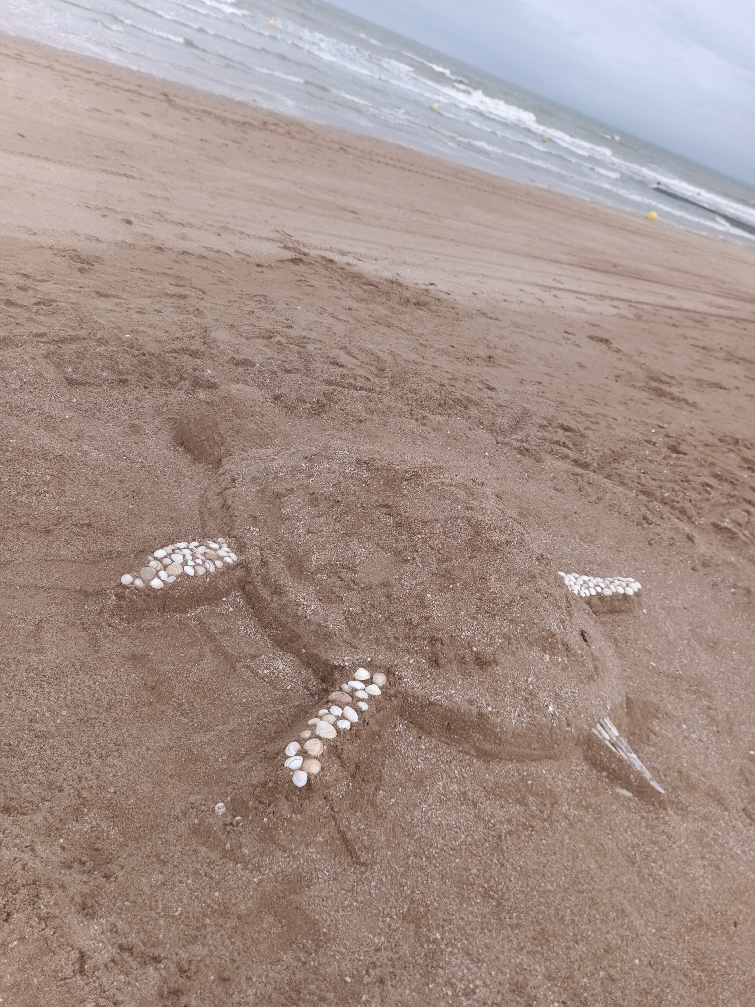 An overhead, oblique shot shows a sand sculpture of a sea turtle on a beach in Houlgate, France. The sculpture is made of wet sand and is decorated with numerous small, white seashells on its flippers and shell. The turtle is positioned in the center of the frame, with its head facing right. The beach extends from the bottom of the frame towards the ocean at the top. The ocean is visible as a light blue expanse with gentle waves breaking near the shore. The sky is overcast and grey. The sand is a light brown color with some debris and footprints visible. A few yellow buoys are scattered in the ocean. The image appears to be taken on a cloudy day.