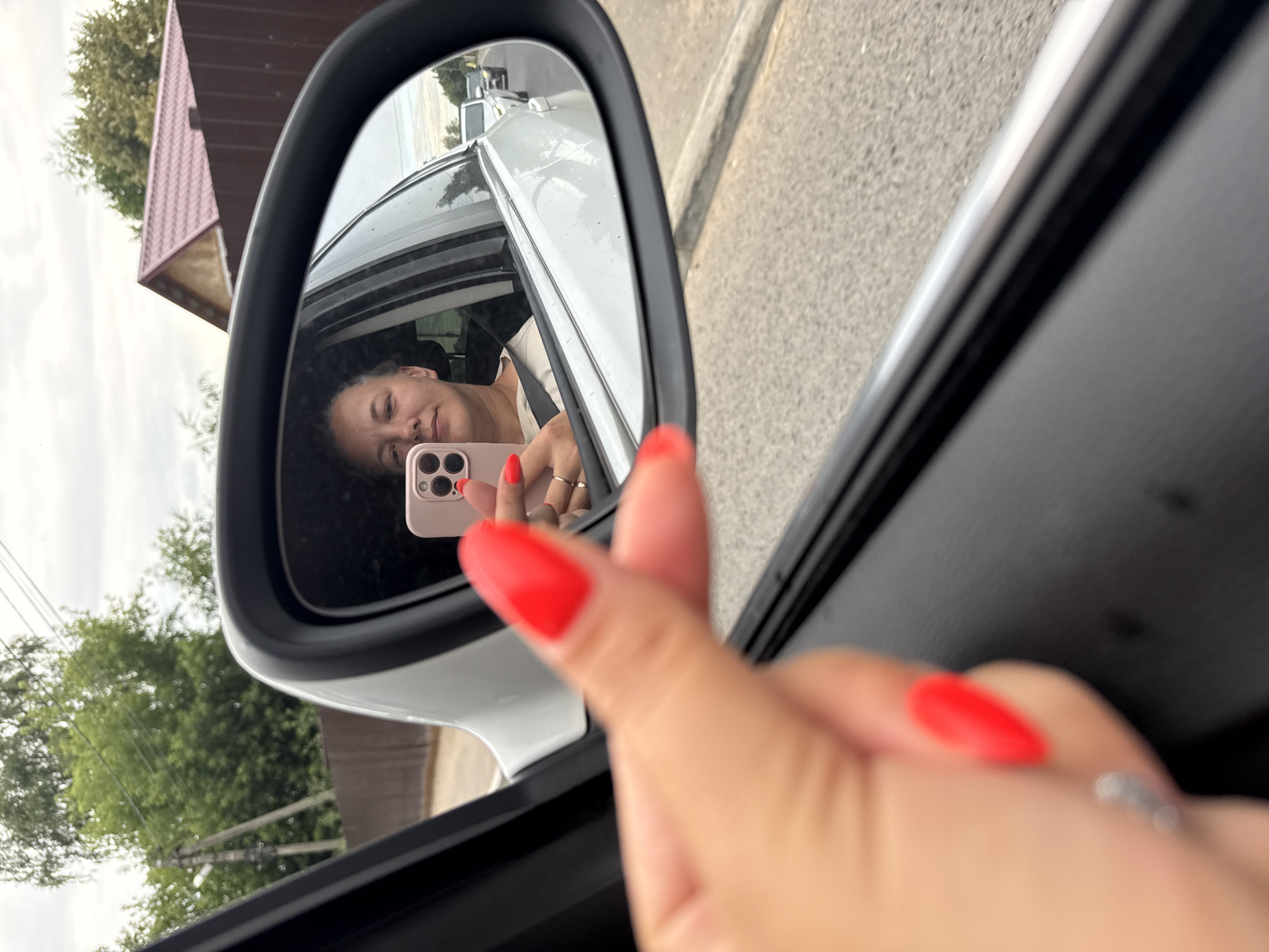 In this media file, a woman is captured taking a selfie in the side mirror of a car. She is sitting in the driver's seat, with her face reflected in the mirror. She is holding a pink smartphone, with the camera facing her. Her nails are painted a vibrant red. In the reflection, she is smiling, and appears to be in good spirits. The background in the mirror shows a street with cars, and some trees and buildings. The exterior of the car is white. The scene appears to be set during the daytime. The location context provided is Dmitrov, Russia. There is no visible text in the image.