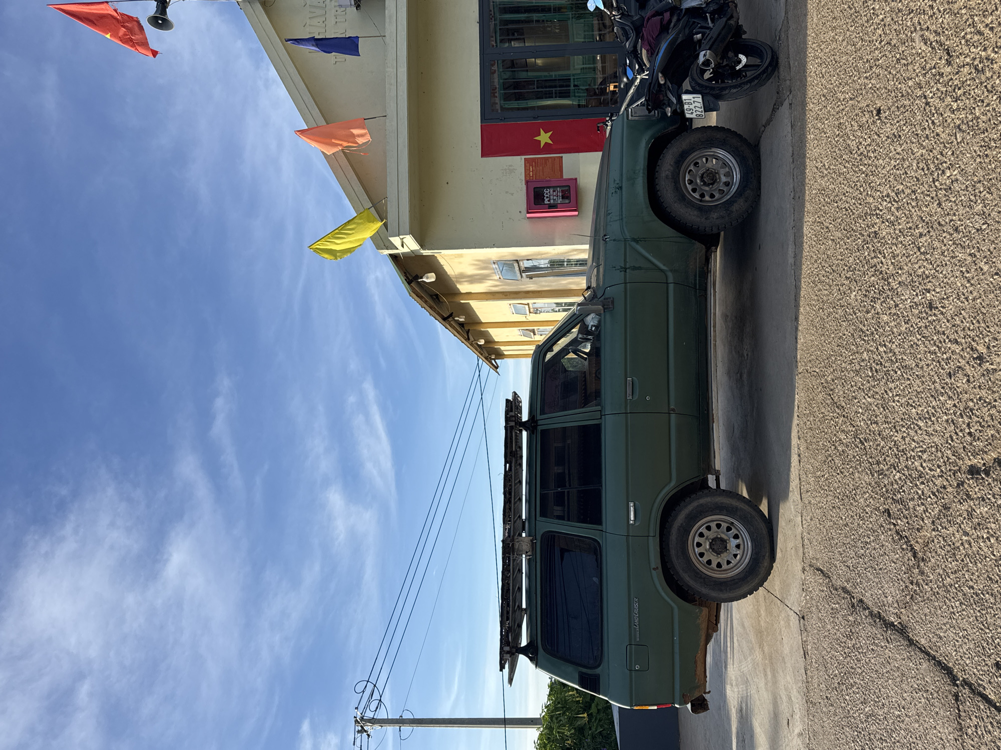 A weathered, olive-green Toyota Land Cruiser is parked on a concrete surface in front of a light yellow building. The vehicle has a roof rack loaded with metal bars. A black motorcycle is parked to the right of the Land Cruiser. The building has a slanted roof with several flags flying, including a red one with a star on the right side, and other colored flags on poles. The sky is blue with scattered white clouds. A utility pole with power lines is visible to the left of the building. The scene appears to be in Trường Thọ, Vietnam, suggested by the text on the building, which includes Vietnamese characters and a license plate on the motorcycle.