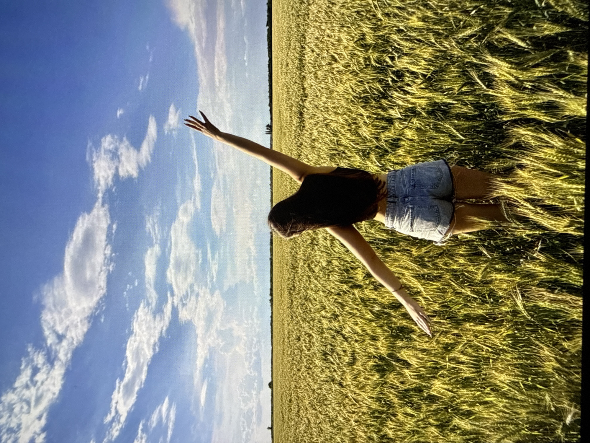 A person is lying down in a field of tall, golden grass under a bright blue sky with scattered white clouds. The person is wearing a dark top and denim shorts. One arm is extended upwards towards the sky, with the fingers spread. The scene appears to be a sunny day, likely late afternoon, judging by the warm light and shadows. The vastness of the field and the open sky suggest a rural or agricultural setting, consistent with a location in Mykolaiv, Ukraine. The person's pose and the overall imagery convey a sense of freedom and connection with nature. No text is visible in the image.