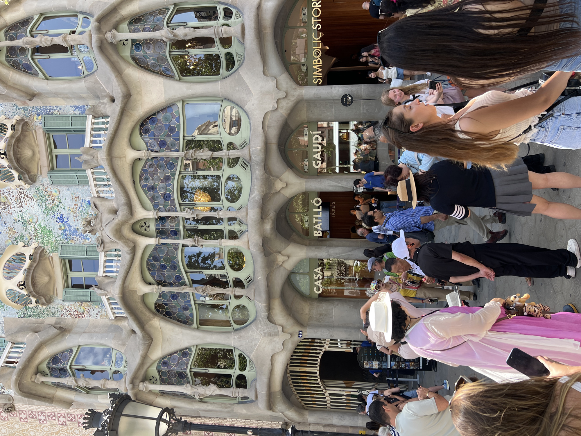 The image shows the exterior of Casa Batlló, a famous building designed by Antoni Gaudí, located on Passeig de Gràcia in Barcelona, Spain. The facade features organic, undulating shapes, colorful mosaics, and uniquely shaped windows, characteristic of Gaudí's architectural style. A crowd of tourists is gathered in front of the building, taking pictures and observing the architecture. Some people are dressed in casual summer clothing, while others are wearing hats. There are signs visible indicating "Casa Batlló" and "Gaudí," as well as a "Symbolic Store." The overall scene depicts a popular tourist attraction with visitors engaged in sightseeing.