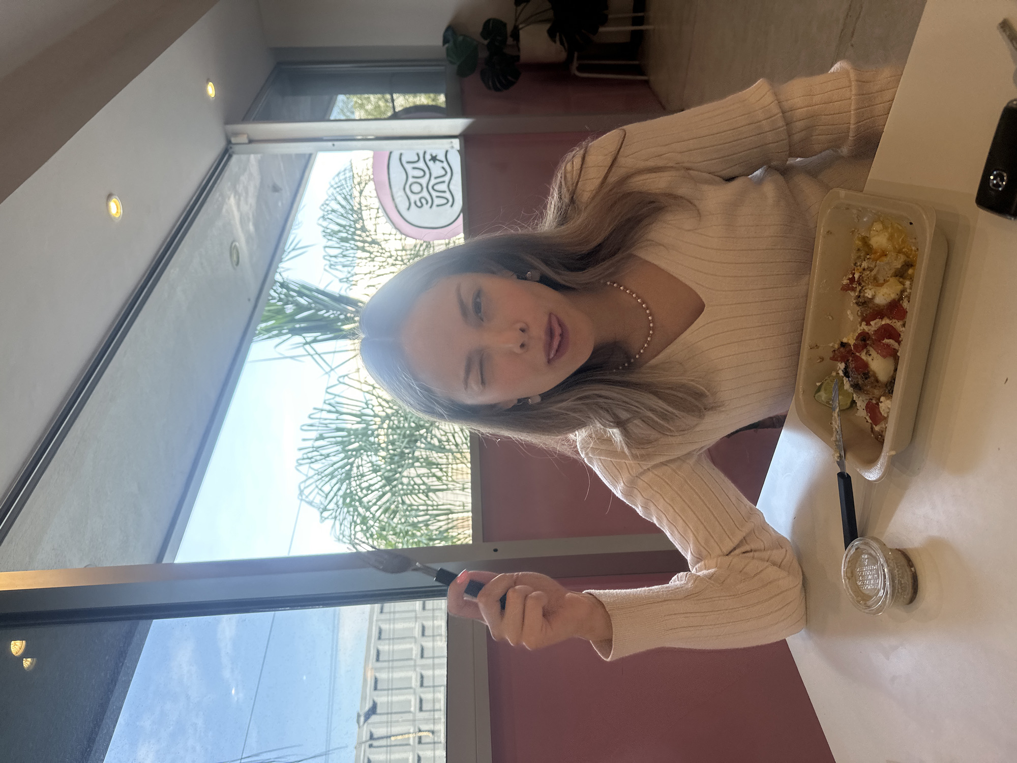 A young woman is photographed at a table, winking and sticking her tongue out. She is holding a fork in her right hand. On the table is a container of food that appears to be partially eaten, as well as a small container. The woman is wearing a light-colored sweater and a pearl necklace. Behind her, a window provides a view of a bright day. Outside the window, palm trees and a building with multiple windows are visible. A sign saying “Soul” is seen in the background. This place is in Puebla, Mexico.