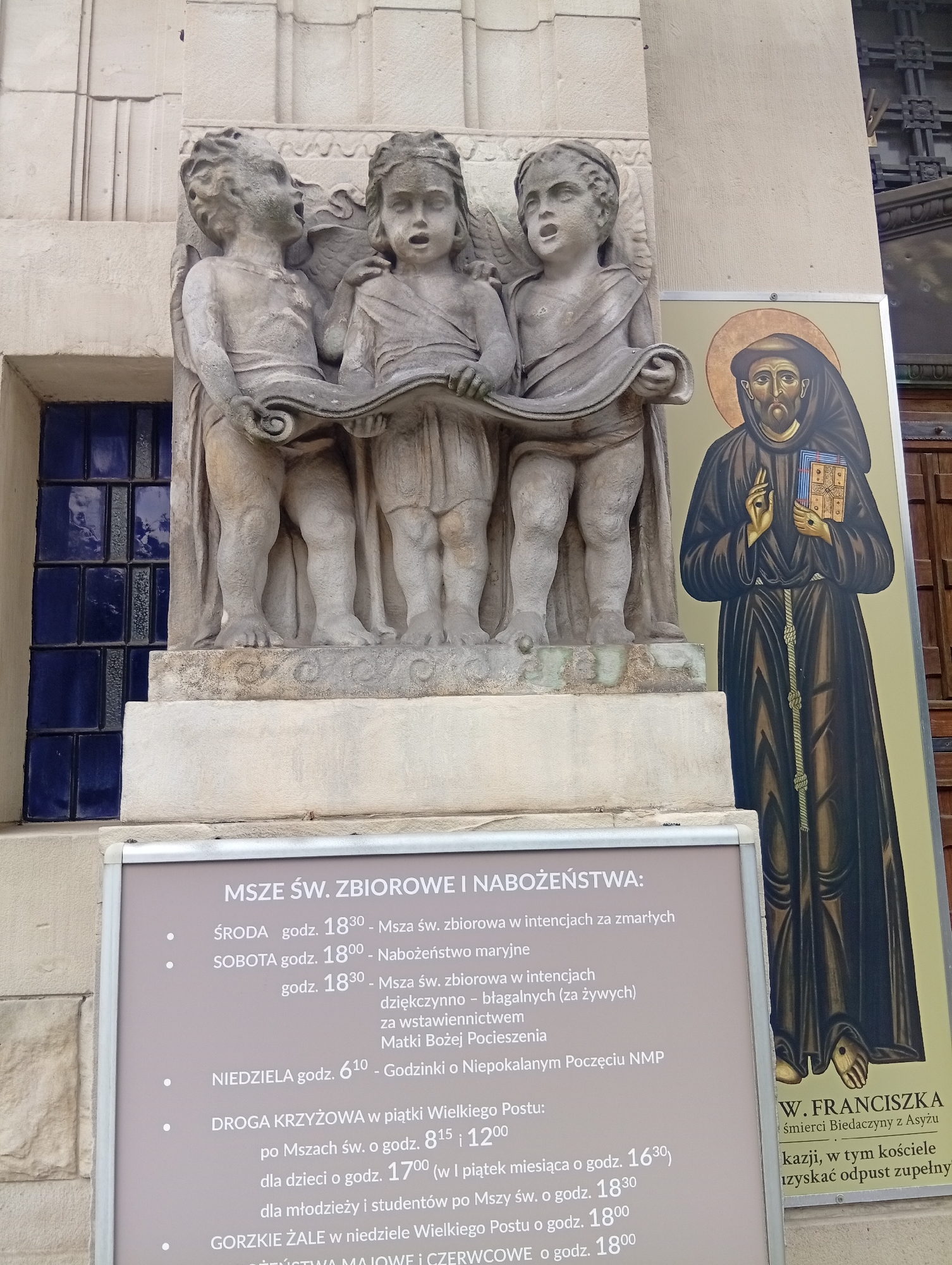 The image shows a facade of a building with a stone sculpture of three cherubs and a painting of a robed monk. A sign with Polish text detailing church services is mounted at the base of the sculpture. The cherubs are depicted holding a scroll. The monk in the painting is wearing a habit and holding a book. The building appears to be made of stone, with a blue-paned window visible to the left of the sculpture. The sign is a light grey color and lists Mass times and other religious observances. The painting to the right of the sculpture features the monk's name, "W. FRANCISZKA," and text in Polish below it. The overall scene suggests a religious institution. The architectural style and the Polish language on the sign indicate the location is likely in Poland.