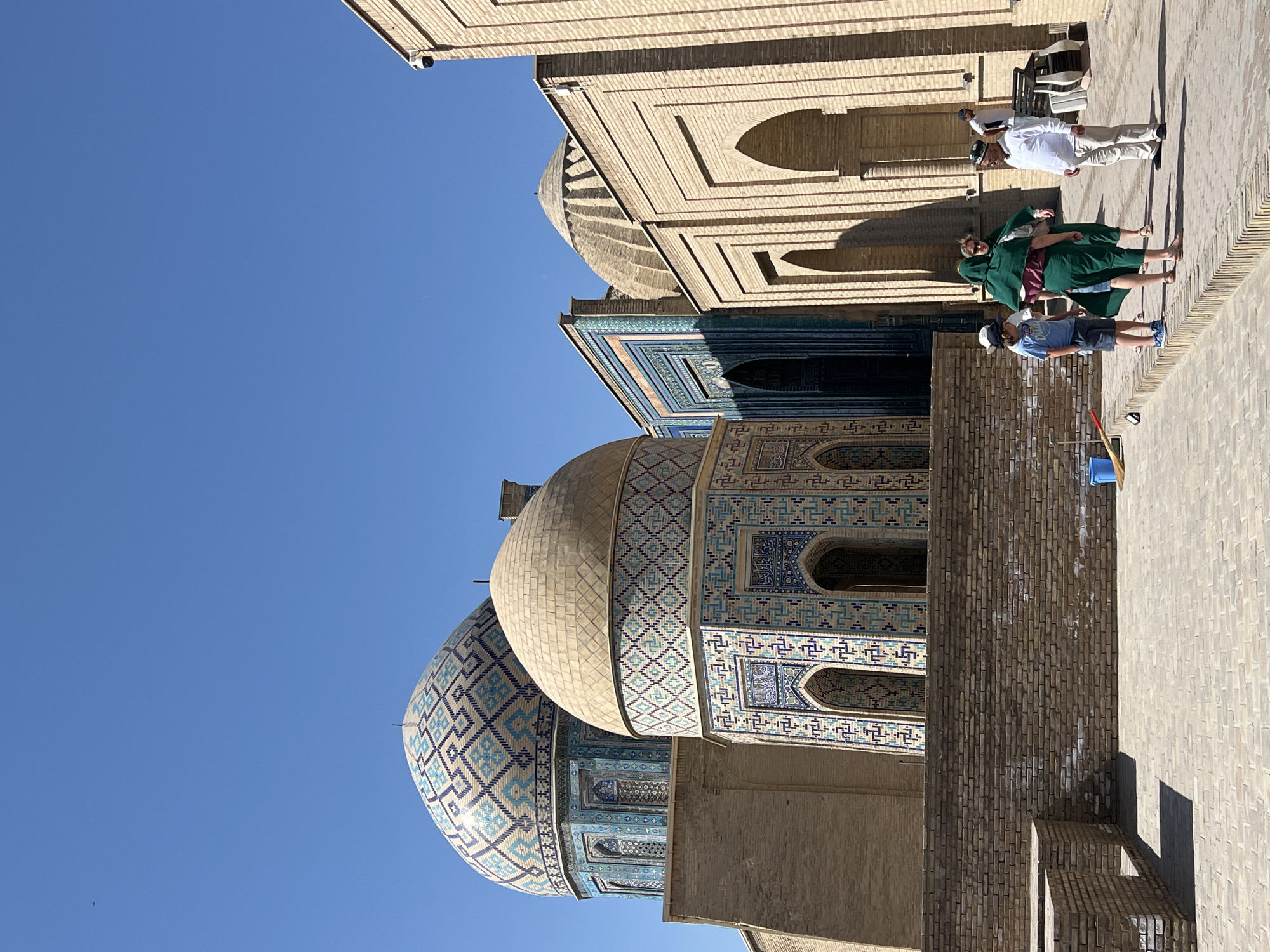 This is a photograph of the Shah-i-Zinda necropolis in Samarkand, Uzbekistan, taken on a sunny day under a clear blue sky. The foreground features a paved courtyard area with a low brick wall. Several people are present: a young boy in a blue t-shirt and shorts, a woman in a green headscarf and dress, and two men, one in a white shirt and light-colored pants, and another in a white shirt and dark pants. They appear to be standing and conversing. In the midground, the iconic blue and turquoise tiled domes and facades of mausoleums are visible, characterized by intricate geometric patterns and arched doorways. The architecture is a striking example of Islamic art and architecture. The background is dominated by the vast expanse of the blue sky, with a single small bird visible in the upper left quadrant. The overall impression is one of historical grandeur and cultural significance.