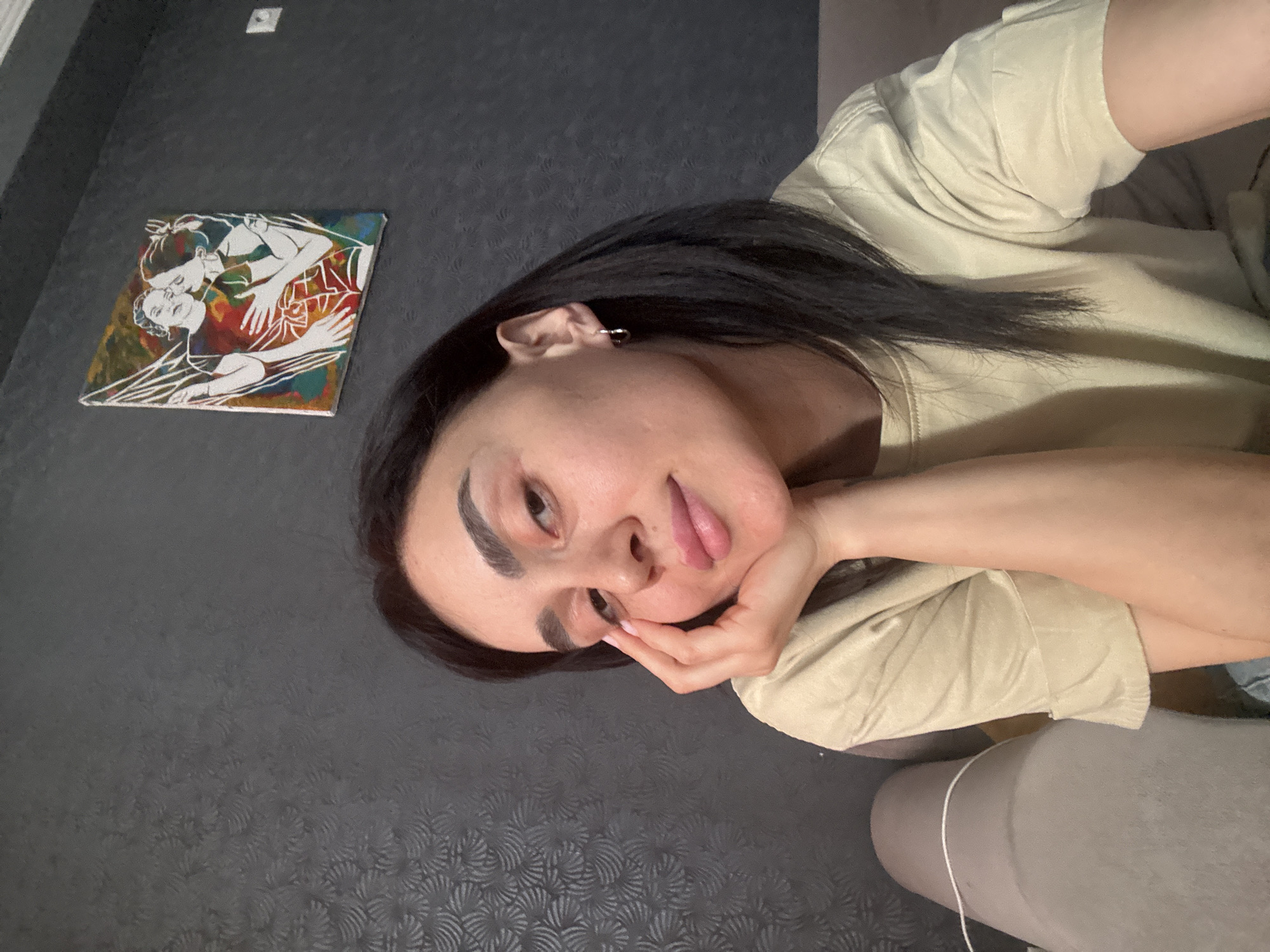 A woman is lying on a couch, posing for a picture. She is wearing a tan-colored t-shirt. The wall behind her is a textured gray. On the wall to her left is a painting depicting two figures in an embrace. A light switch is located above the painting. The location appears to be indoors, possibly a residence. There is no indication of the location being in Tbilisi, Georgia, from the image.