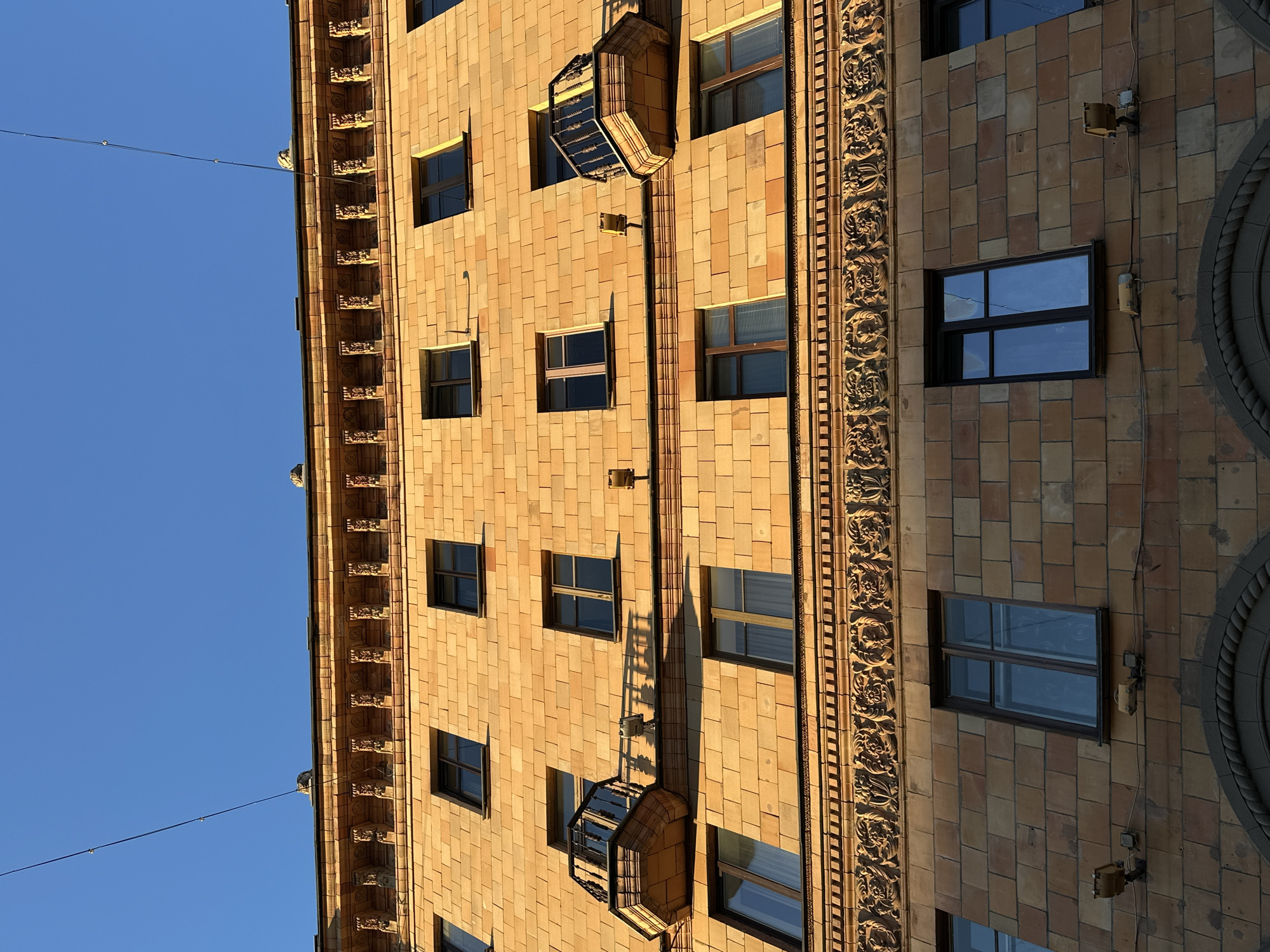 The image displays the facade of a building, likely an apartment building or hotel, with light brown, textured tiles. The building features numerous windows with dark frames, many reflecting a clear blue sky. Several decorative elements are visible, including ornate carved trim running vertically along the building and protruding window boxes with metal detailing. Light fixtures are attached to the exterior walls. The orientation of the image suggests it was taken from below, looking upwards at the building. The clear sky and warm lighting indicate it is daytime, likely late afternoon or early morning due to the long shadows cast by the protruding architectural features. The image is oriented vertically. No people or specific activities are depicted, focusing solely on the architectural details of the building. The location context is provided as Minsk, Belarus.
