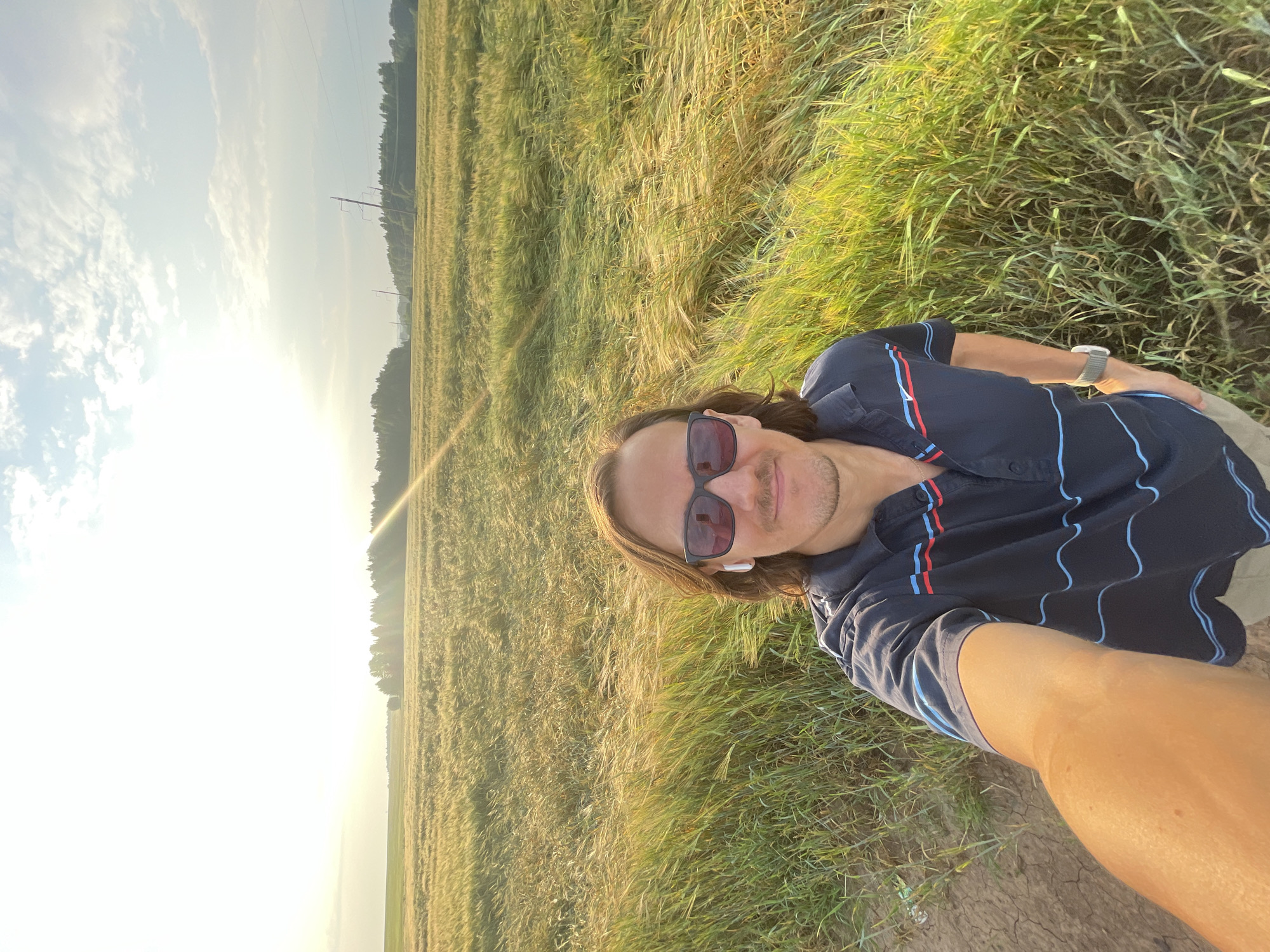 In the city of Ol'khovo, Russia, a selfie is taken in a field of tall, golden-green grass under a bright, partly cloudy sky. The sun, low on the horizon to the left, casts a warm glow and a lens flare across the scene. A man with long brown hair and sunglasses is centered in the frame, facing the camera. He is wearing a dark blue collared shirt with thin red and blue horizontal stripes, light-colored shorts, a white smartwatch on his left wrist, and white wireless earbuds. The field extends into the distance, with trees and power lines visible along the horizon. The ground in the foreground to the left appears to be dry, cracked earth.