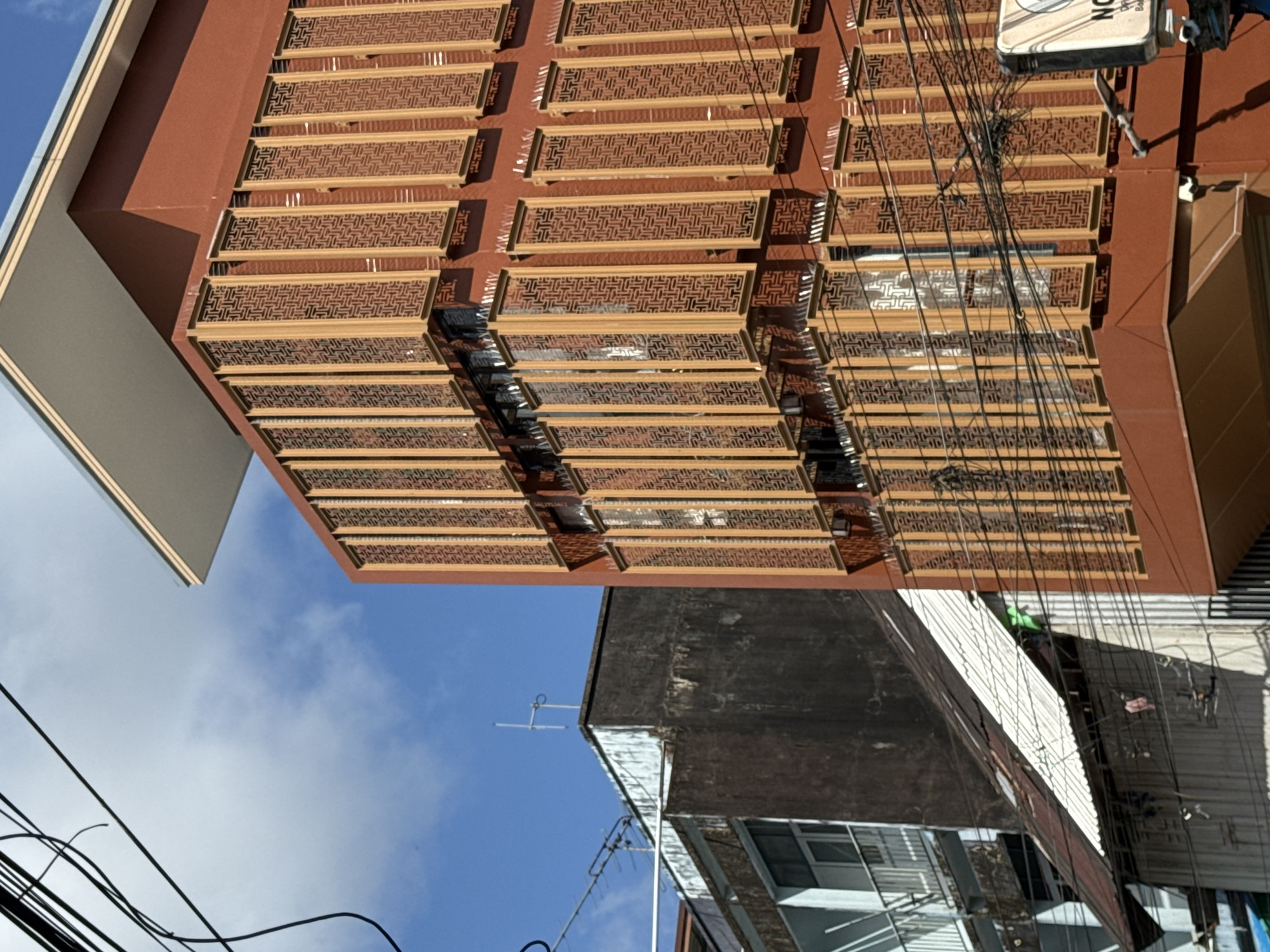 The image captures an urban street scene in Ranong, Thailand, on a clear day with a blue sky and scattered clouds. The foreground features the side of a modern, reddish-brown building with decorative, latticed screens covering its windows. These screens are composed of vertical wooden or metal panels with an intricate, repeating geometric pattern. The building has a clean, angular design with a white overhang.
To the left of this modern structure, an older, darker building with a weathered facade is visible. This older building has multiple stories with balconies and windows, some appearing to have barred railings. A corrugated metal roof, partially rusted and white, slopes downwards from the older building towards the foreground, partially obscuring the lower levels of the buildings. Overhead, a complex network of black electrical wires crisscrosses the sky, suggesting an urban environment with overhead power lines.
In the bottom right corner of the image, a white sign with black lettering is partially visible, indicating a business or establishment. The overall impression is one of a densely built urban area where new architectural styles are juxtaposed with older, more utilitarian structures. There are no people visible in the frame. The lighting suggests it is daytime, with clear sunlight casting shadows.