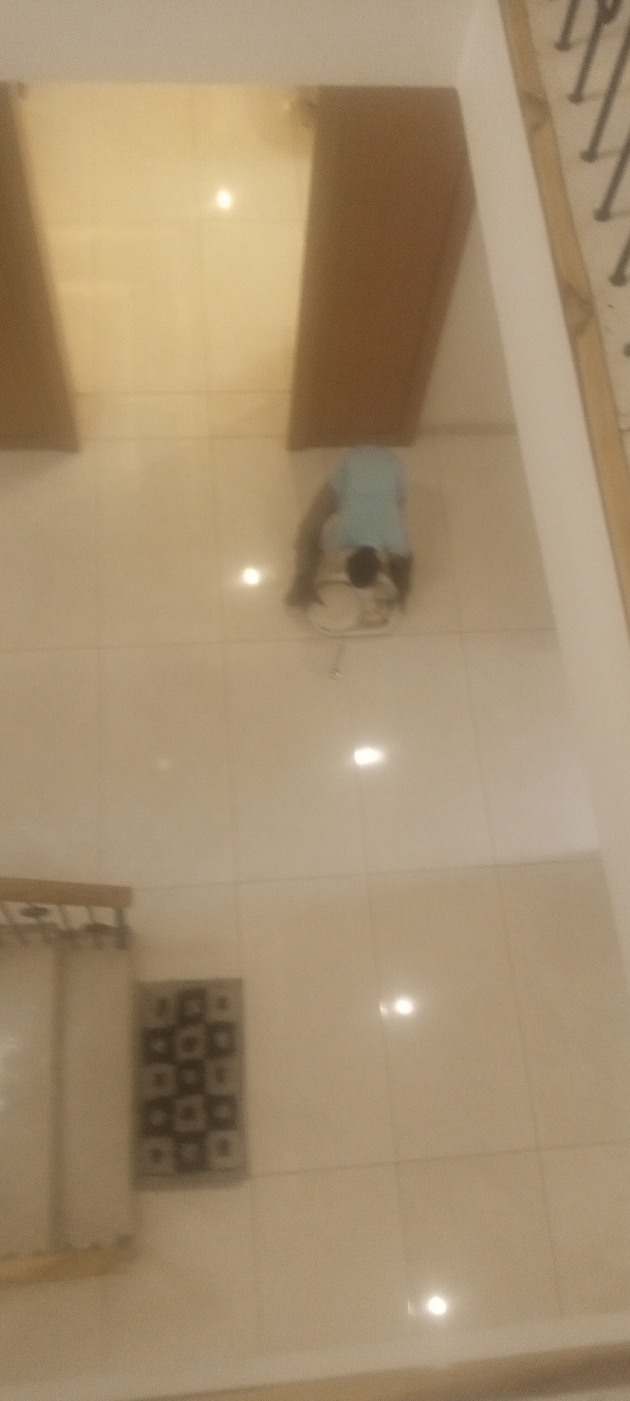 A person is kneeling and cleaning the floor in what appears to be an indoor tiled space in Unguwar Rimi, Nigeria. The space contains two brown wooden doors on the upper portion of the image. A dark patterned rug and a partially visible piece of furniture are located at the bottom left of the image. A section of staircase railing is visible on the right.