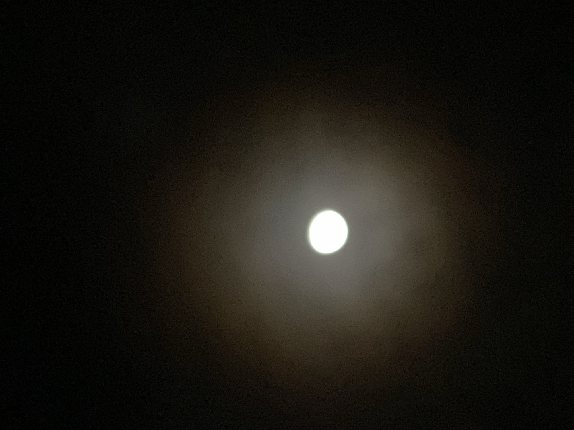The image shows a bright, full moon centered in the dark night sky. There is a hazy or diffused ring of light around the moon, possibly due to atmospheric conditions. The surrounding sky is dark, with no other visible stars or objects. No specific information about the location in Maigizo, Nigeria can be determined from the image. There are no people, objects, or actions depicted other than the presence of the moon in the night sky.
