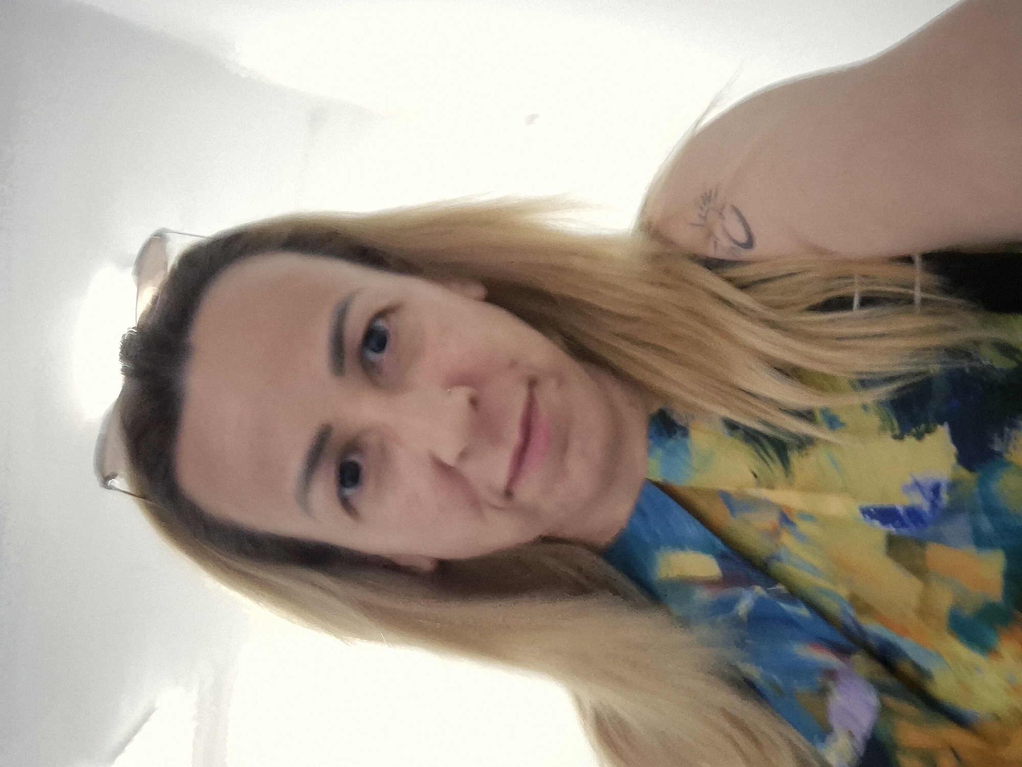 This is a selfie of a person with blonde hair. The image is oriented sideways. The person is looking at the camera with a slight smile. They are wearing a colorful, abstract patterned shirt with yellow, blue, and green colors. A tattoo is visible on their upper arm. The background is a plain, light-colored wall, suggesting an indoor setting. The lighting is bright, possibly from natural light. No text is visible in the image.