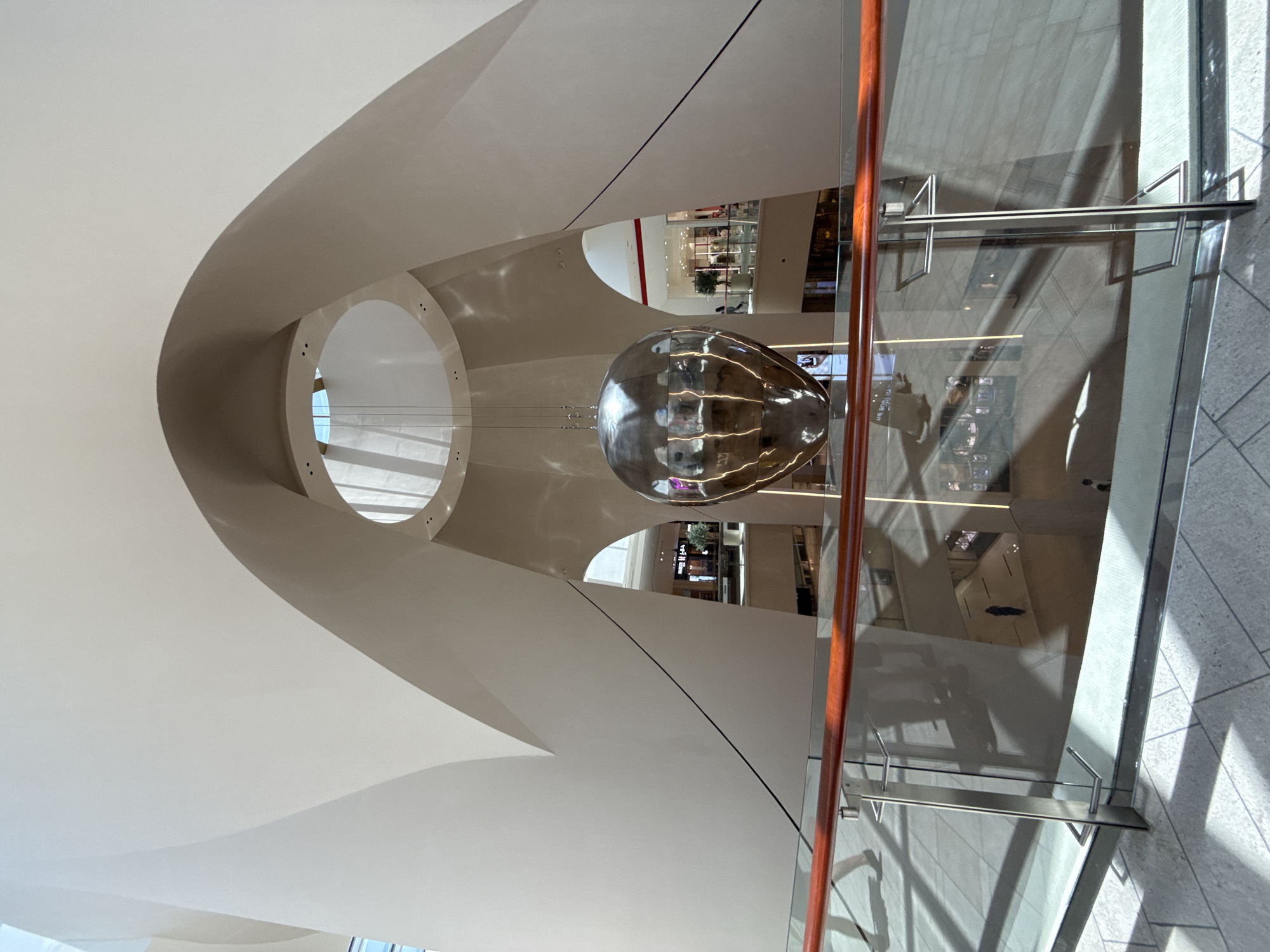 This image captures a view from the upper level of a contemporary shopping mall, likely in Dubai, United Arab Emirates, with its distinctive modern architecture and expansive interior. Dominating the center of the frame is a large, reflective, spherical sculpture suspended from the ceiling by thin wires. The sculpture's polished surface mirrors the surrounding architecture and the light filtering in from above.
The mall's interior features smooth, curved white walls that form a dramatic atrium. A circular opening in the ceiling allows natural light to illuminate the space. Below, the image shows multiple levels of the mall, with shops visible through archways and glass railings. The floor is a light-colored, tiled surface, with geometric patterns of light and shadow cast by the sunlight.
A glass and wood railing separates the viewer from the lower levels, with reflections of people and the environment visible on the glass. Two figures are visible walking on the upper level, their forms partially obscured by the railing. On the lower levels, several individuals are seen walking, their presence indicated by their silhouettes and reflections. The overall impression is one of a spacious, well-lit, and architecturally striking public space.