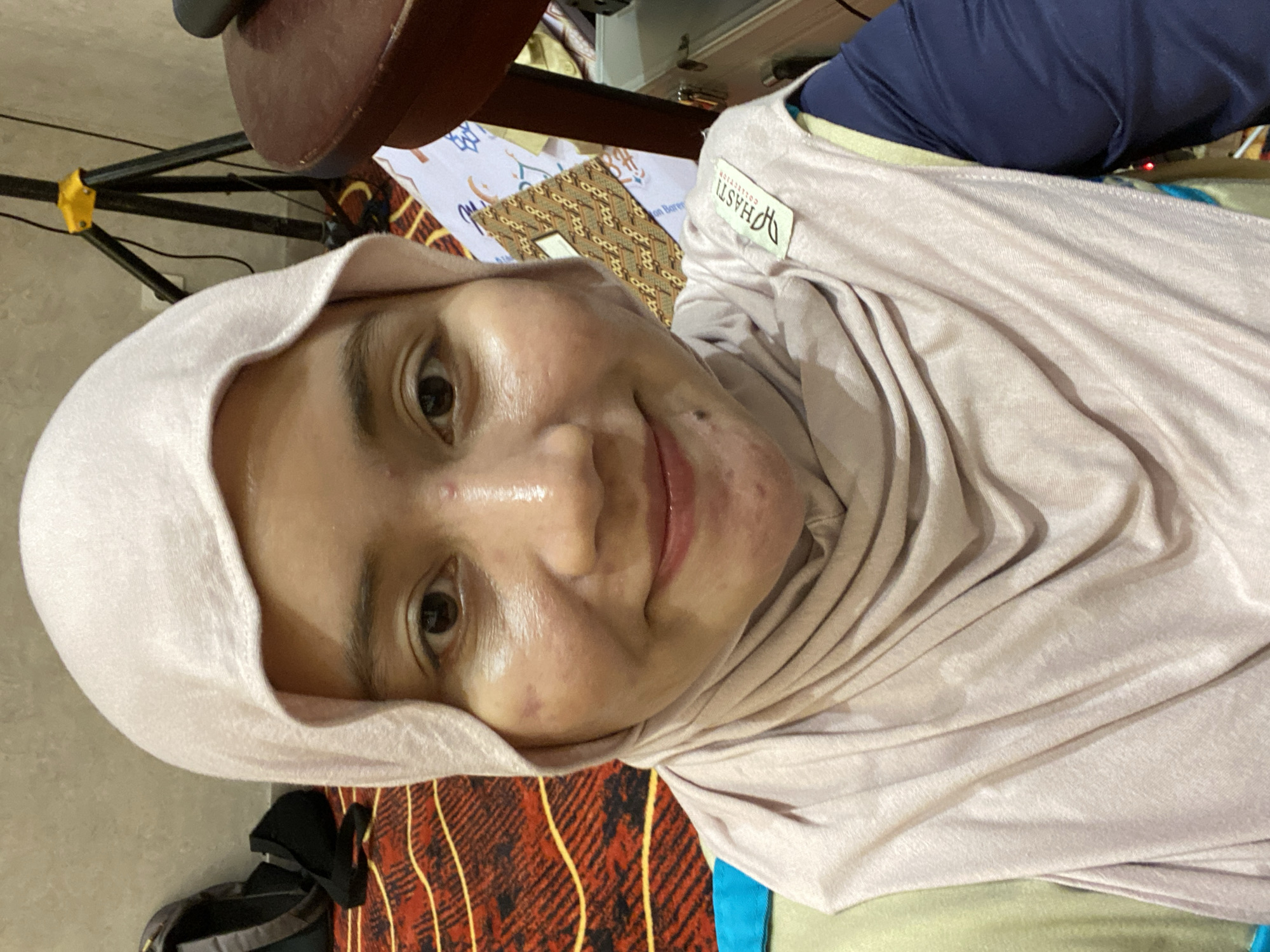 A selfie shows a woman wearing a light pink hijab and a matching scarf. She is smiling and looking at the camera. Her face has some visible blemishes. Behind her, a patterned rug is visible on the floor, along with a black backpack and a tripod. A dark wooden table is to her right, with some papers and other objects on it. The overall lighting suggests an indoor setting. The specific location in Banjarbaru, Indonesia, cannot be determined from the image alone.