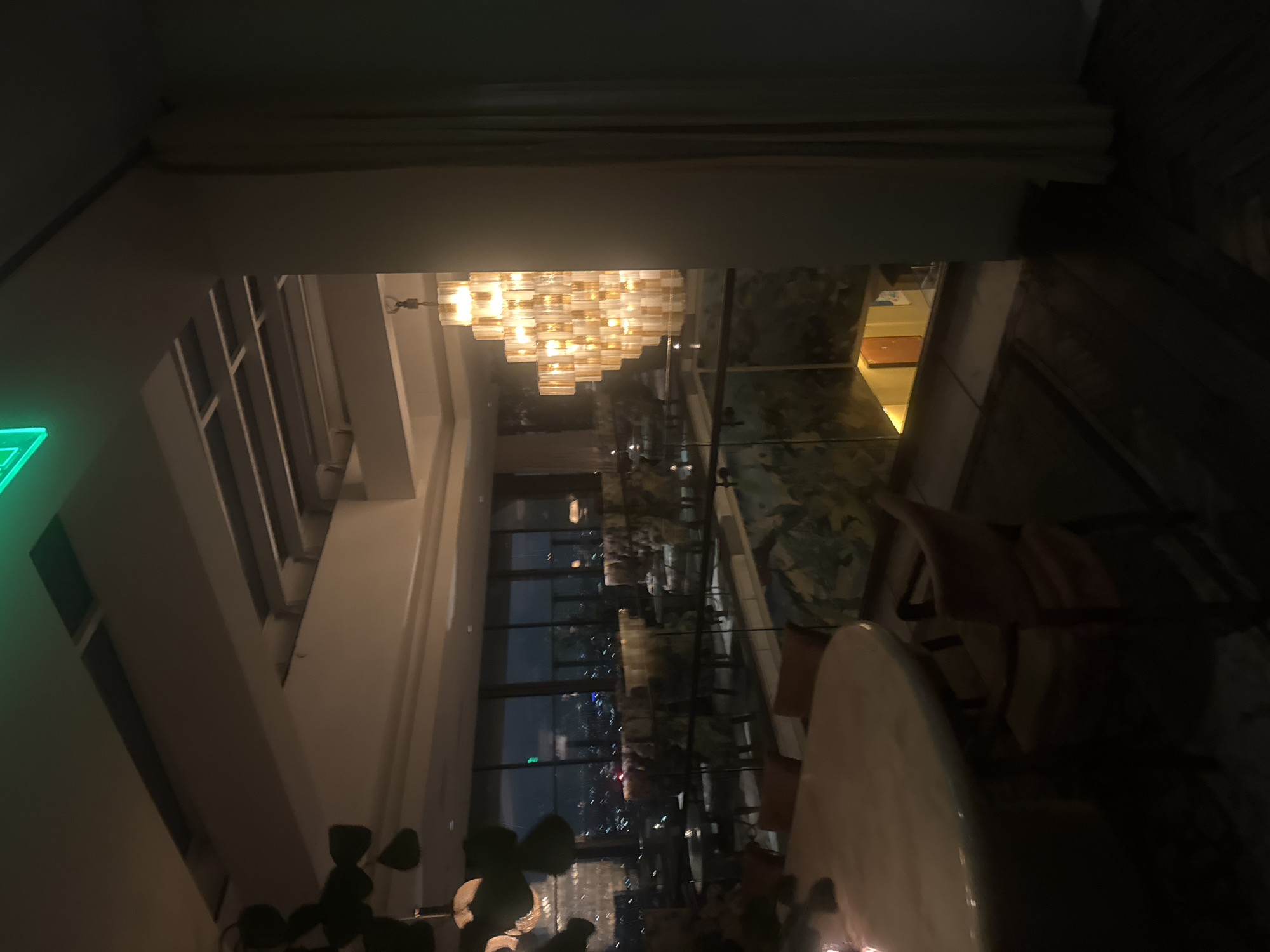 The image shows an interior view of a restaurant or bar located in West Hollywood, California, United States of America, likely at night. A large, multi-tiered chandelier dominates the upper-center of the frame. A vertical panoramic window provides a view of the city at night. Tables and chairs are visible closer to the bottom of the frame, indicating a dining or seating area. A partially visible green exit sign is present on the far left. The overall lighting is dim.