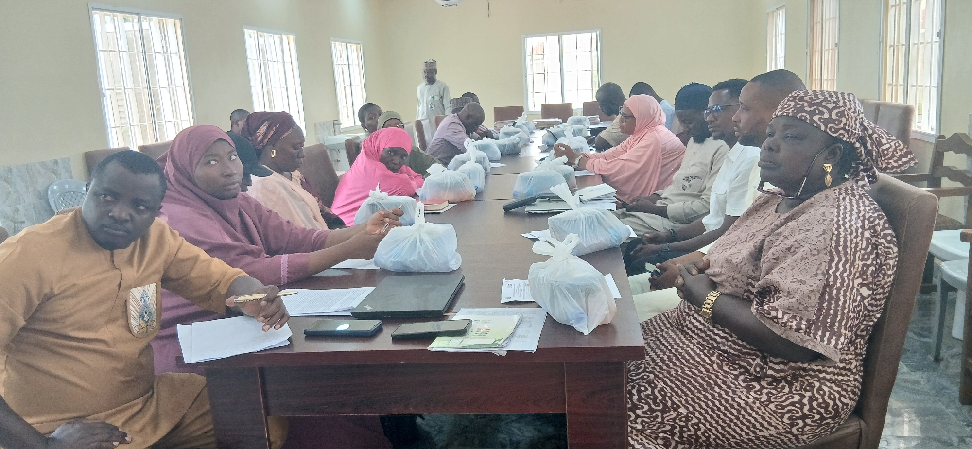 The image shows a group of people, likely attending a meeting or training session, seated around a long table in a room. The subjects are diverse in age and gender, with many wearing traditional African attire, including headscarves and long robes. Several individuals are looking towards the camera or engaging with the person taking the photograph.
On the table, there are several white plastic bags, possibly containing food or materials, along with laptops, mobile phones, papers, and pens. This suggests a formal gathering where participants may be receiving or distributing items, or engaging in work-related activities.
The setting appears to be an indoor meeting room. The walls are painted a light color, and large windows with metal bars are visible, suggesting a potentially secure or utilitarian building. The overall lighting is bright, indicating it is daytime. The presence of a whiteboard or projection screen is suggested by a faint outline on the wall in the background, and a person standing in the background wearing a traditional cap suggests this is likely in Nigeria.
The mood seems to be one of attentiveness and engagement, with some individuals appearing serious and focused, while others have a more relaxed or curious expression. The presence of multiple people in a meeting setting indicates a social or professional event. No specific text is clearly legible in the image.