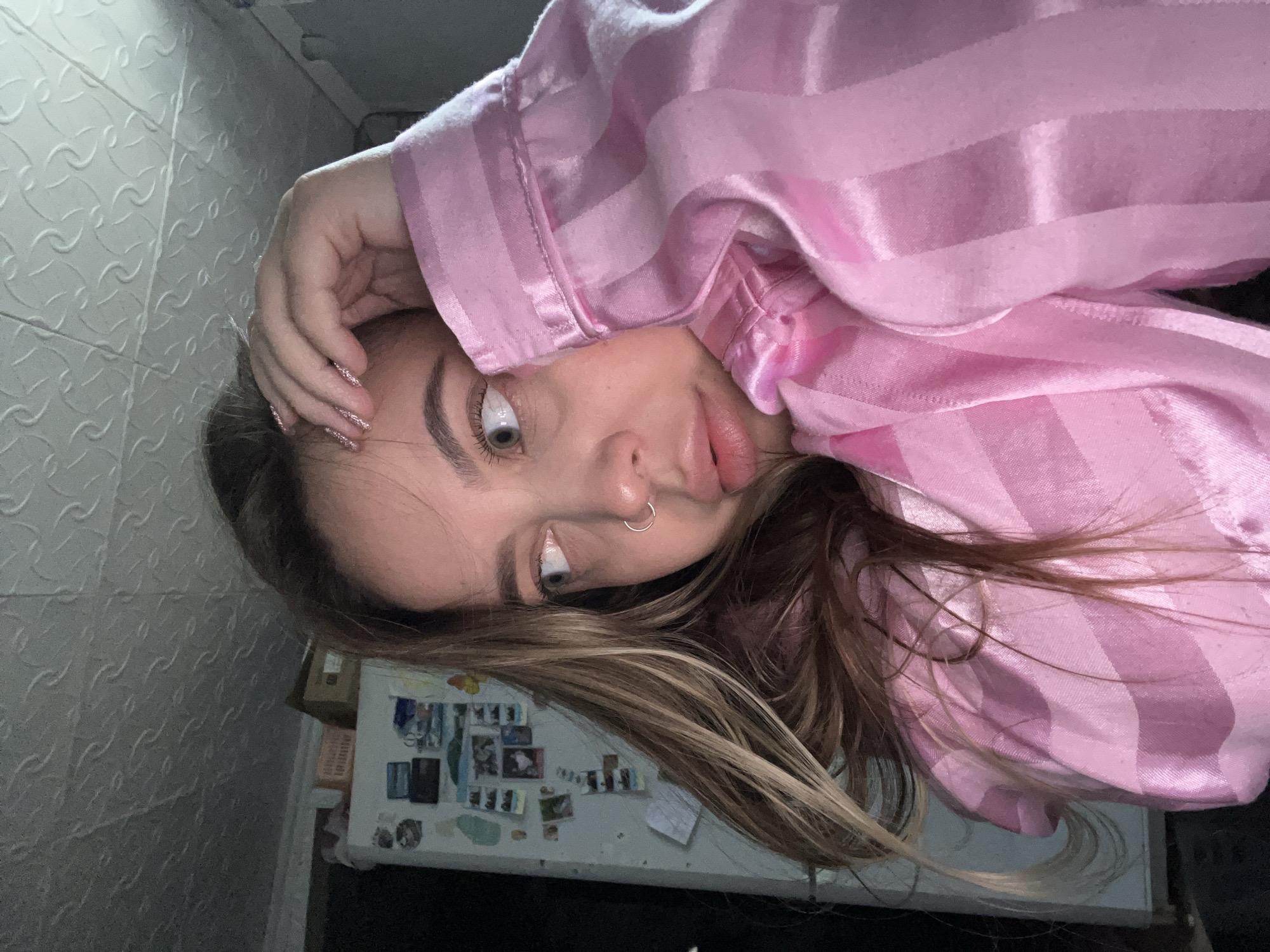 A young woman with light brown hair, light eyes, and a nose ring is photographed in a close-up shot. She is wearing a pink striped pajama top. Her hand is resting on her forehead. Behind her is a wall and what appears to be a refrigerator covered with photos and notes. There is no indication that this image was taken in Novopokrovskaya, Russia.