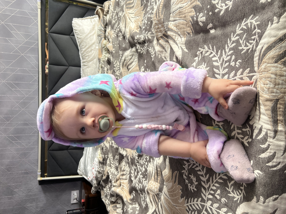 The image displays an infant seated upright on a bed within an indoor room in Leninakan, Russia. The infant wears a multi-colored plush hooded onesie in shades of lavender, light blue, pink, and yellow, adorned with light-colored star patterns. A grey pacifier with a pink nipple is held in the infant's mouth, connected by a pink beaded pacifier clip featuring white and pink beads, some forming letters, which attaches to the onesie. The infant has light-colored hair, visible ears with small hoop earrings, and blue eyes directed upwards. Light pink socks are worn on the infant's feet, with hands resting on or near the feet.
The bed features a dark grey padded headboard with geometric paneled sections. It is covered by a dark grey blanket with an intricate light-colored leaf and floral pattern. A single cream or white pillow is visible against the headboard on the right side. To the left of the bed, a dark wooden nightstand is partially visible, displaying a white power outlet and a dark cylindrical object. The wall behind the bed is covered with a grey wallpaper featuring a subtle geometric pattern.