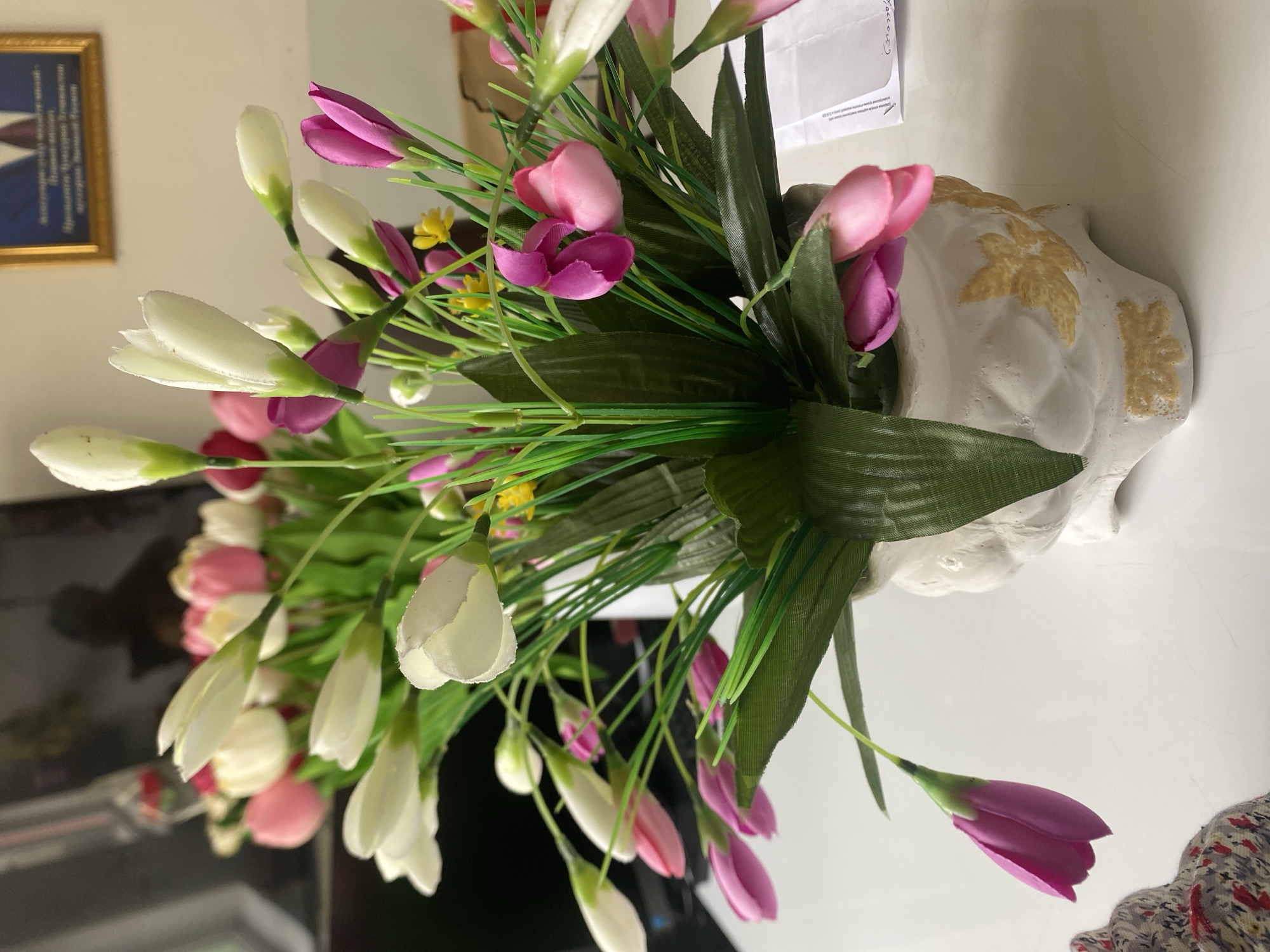 A close-up, eye-level shot features a white ceramic vase containing a bouquet of artificial flowers. The vase is intricately designed with raised, golden floral patterns and sits on a white, reflective surface, likely a table. The bouquet is comprised of various artificial flowers, predominantly white and pink tulip-shaped buds, interspersed with green foliage and a few small yellow accents. The background is blurred but suggests an indoor setting, with a dark rectangular object, possibly a monitor or television, on the left, and a framed picture on the wall to the right. The overall lighting is bright, casting subtle shadows. The scene is devoid of any human presence. Based on the framed picture, which appears to contain official symbols or portraits, and the general ambiance, the location is likely an office or a formal room within the city of Dushanbe, Tajikistan.
