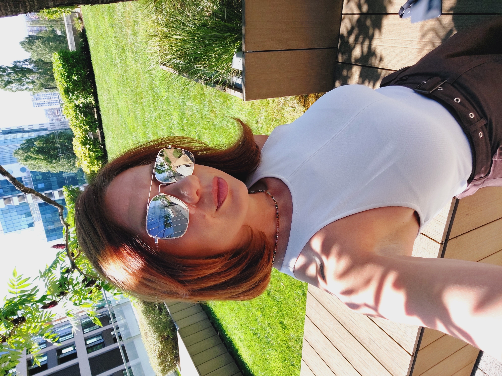 A woman with auburn hair and sunglasses is taking a selfie outdoors in a city setting. She is wearing a white sleeveless top and a brown skirt. The background shows a green lawn, some bushes, trees, and modern buildings. The scene appears to be on a rooftop terrace or a elevated urban park. The lighting suggests it is daytime. The reflection in her sunglasses shows a street with buildings and trees.
