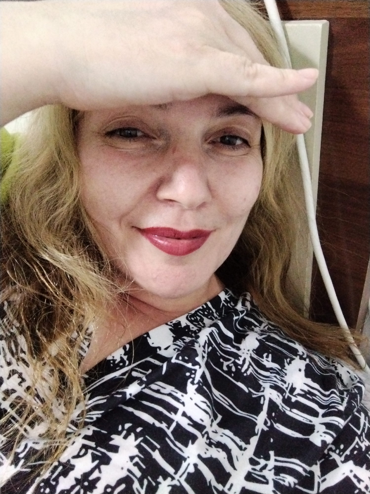 A woman is posing for a selfie indoors. She has fair skin, light brown hair, and is wearing dark red lipstick. Her hand is raised to her forehead. She is wearing a black and white patterned top. A white cord runs vertically along the right side of the frame, next to a beige wall. The environment appears to be a room within a residence, given the plain wall and generic decor. There is no specific evidence to pinpoint the location to Al Mas'oudi, United Arab Emirates, without additional information.