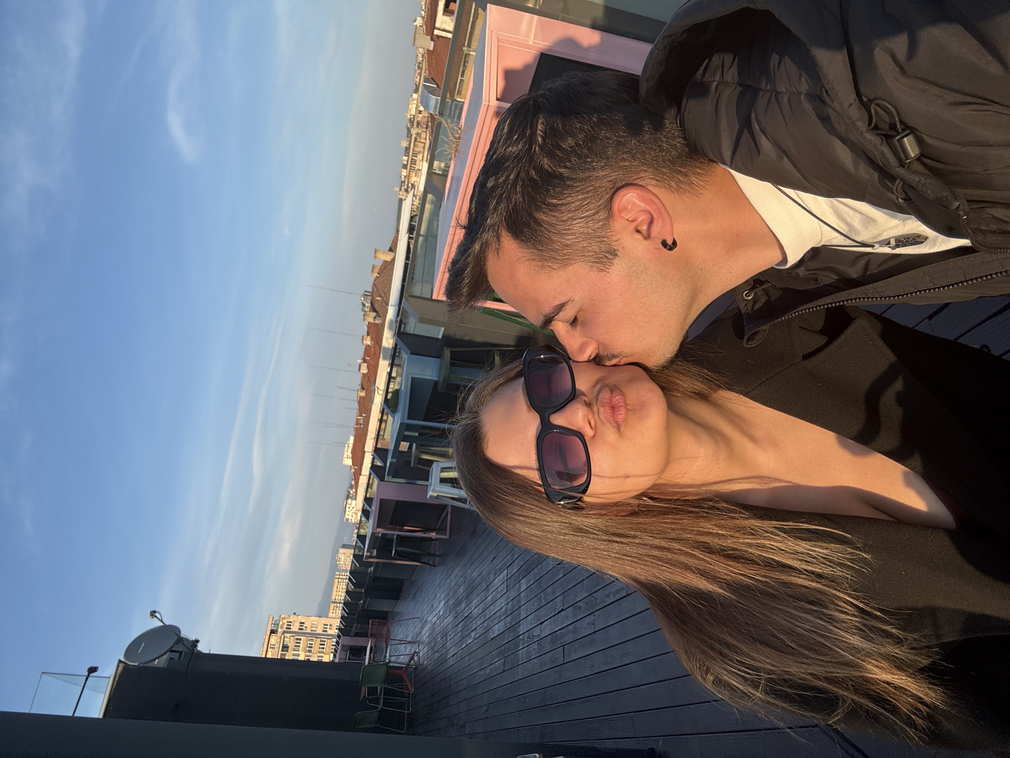 A couple, a man and a woman, are posing for a selfie on a rooftop in Sofia, Bulgaria. The man is kissing the woman on the cheek. The woman is wearing sunglasses and has her lips pursed. The rooftop is made of wooden planks and has some chairs and tables. In the background, there are buildings and a clear blue sky.