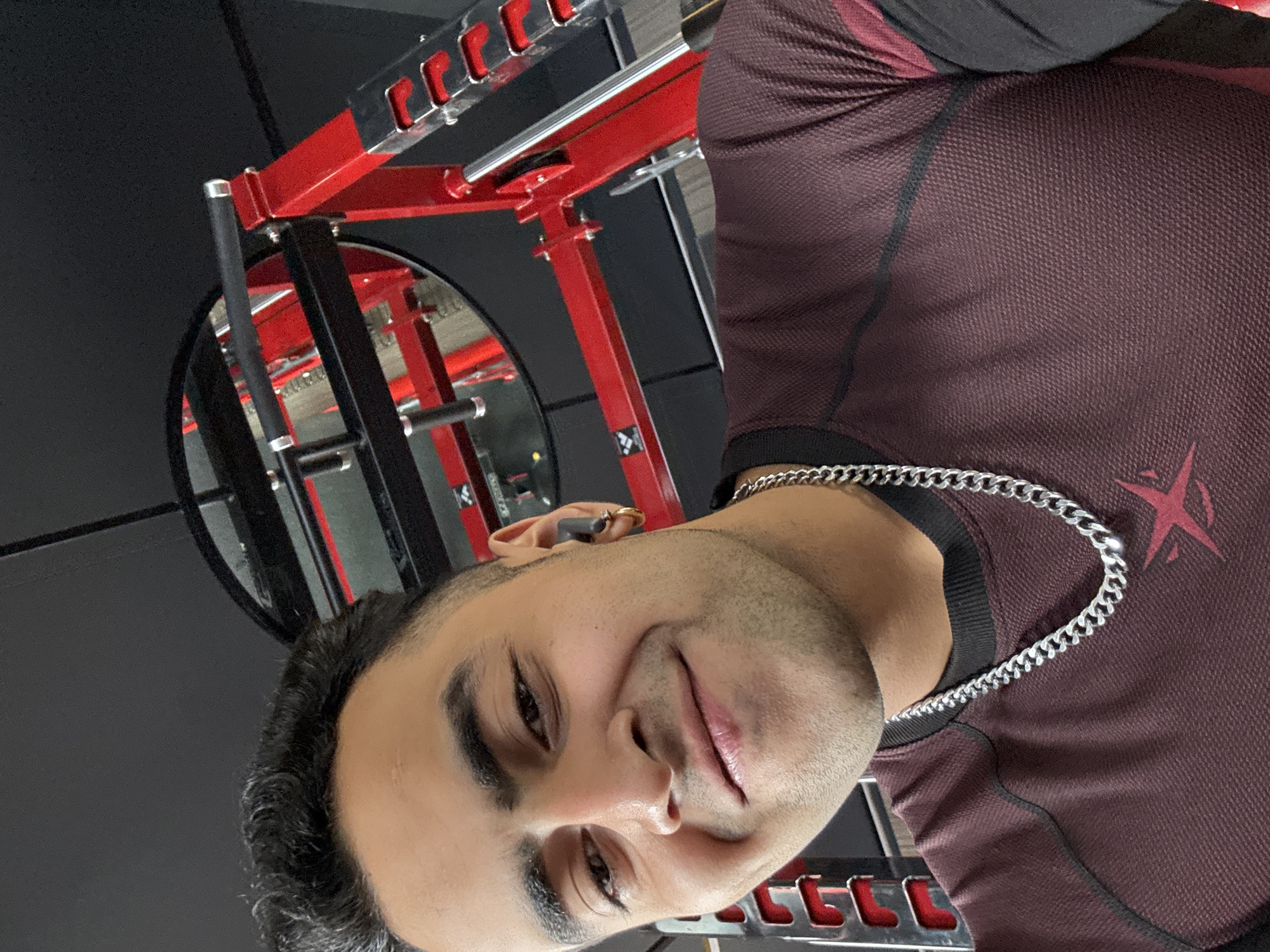 A close-up selfie shows a man with dark hair, a short beard, and a silver chain necklace. He is wearing a maroon and black long-sleeved athletic shirt with a red emblem on the chest. He has a slight smile and is looking at the camera. The background appears to be a gym, with a red squat rack and a mirror reflecting the equipment. The image is not located in Kolkata, India.