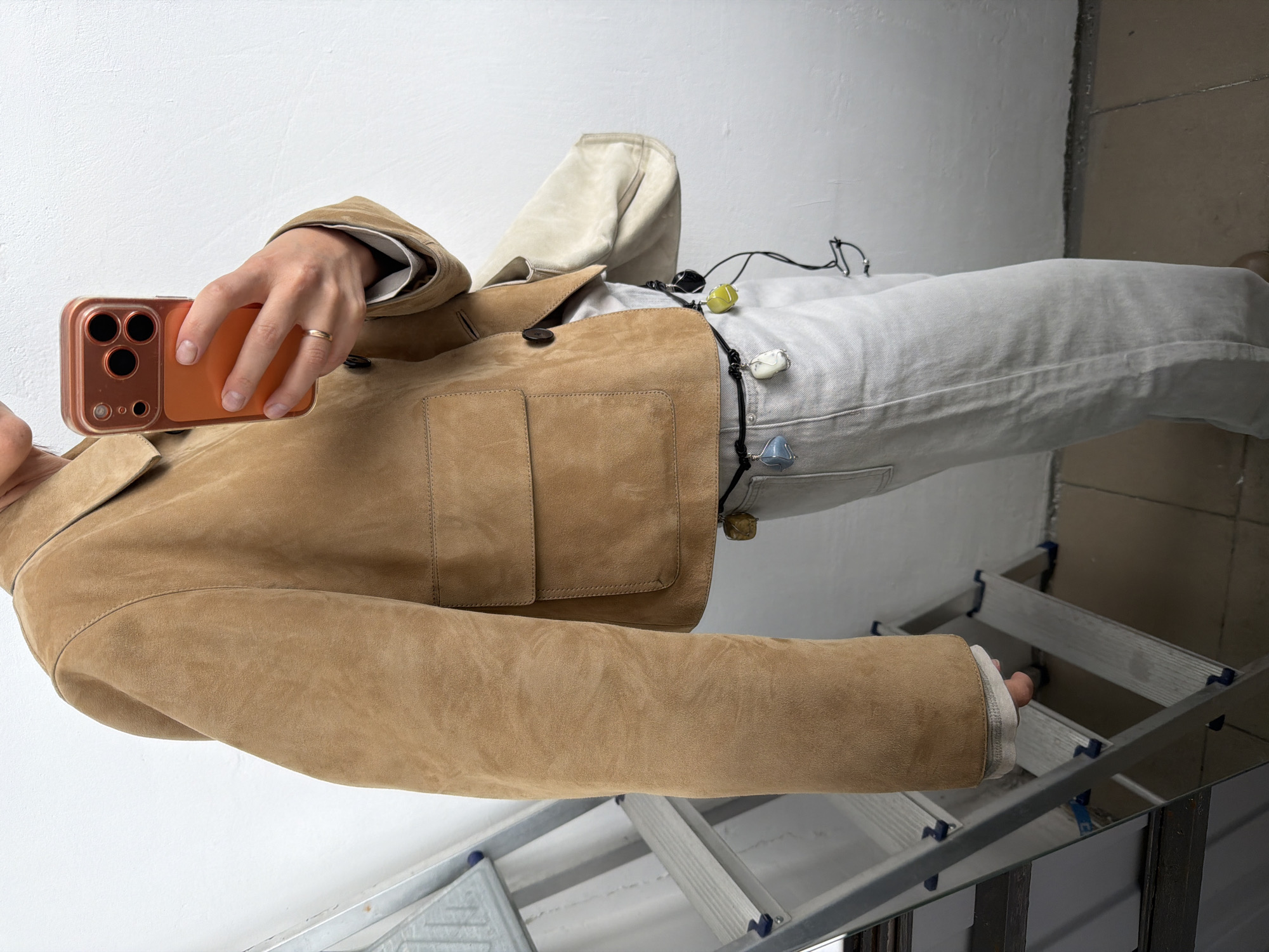 A person stands on a ladder indoors, in a room with white walls. They are holding an orange smartphone in their right hand, taking a picture. The person is wearing a tan suede jacket and light-colored denim jeans. A decorative necklace with colorful stones hangs from the waist of the jeans. The lower portion of the ladder is visible beneath the person. The location is not discernible as Khimki, Russia from this image.