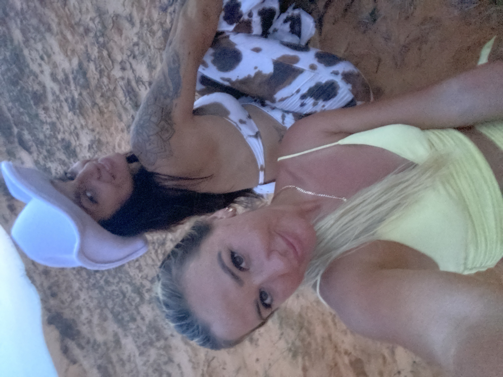 Two women are sitting in a sandy environment. The woman in the foreground, on the left, has blonde hair and is wearing a light yellow top. She is looking towards the camera, smiling. The woman in the background, on the right, is wearing a white cowboy hat and a bikini with a cow print pattern. She is also looking towards the camera and smiling. The background is a sandy hill. The scene appears to be outdoors, likely in a desert or beach-like setting. While the specific location of Mui Ne, Vietnam, is mentioned in the prompt, there are no visual cues in the image to confirm this.