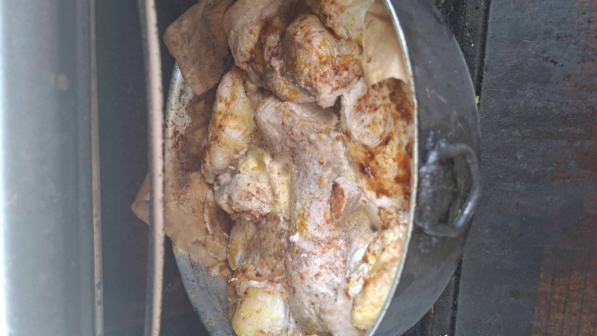 A close-up, overhead shot shows a large, round metal pot filled with pieces of seasoned meat and parchment paper, cooking inside what appears to be an oven or smoker. The meat, likely chicken or pork, is pale with a dusting of reddish-orange spices. The parchment paper is folded around the meat, creating a rustic presentation. The pot has two looped handles and is resting on a dark, textured surface, possibly a grill or a stone hearth. The background is dark and out of focus, with visible metal racks suggesting an enclosed cooking environment. The scene is devoid of people or specific locations that would indicate Voinova, Moldova.
