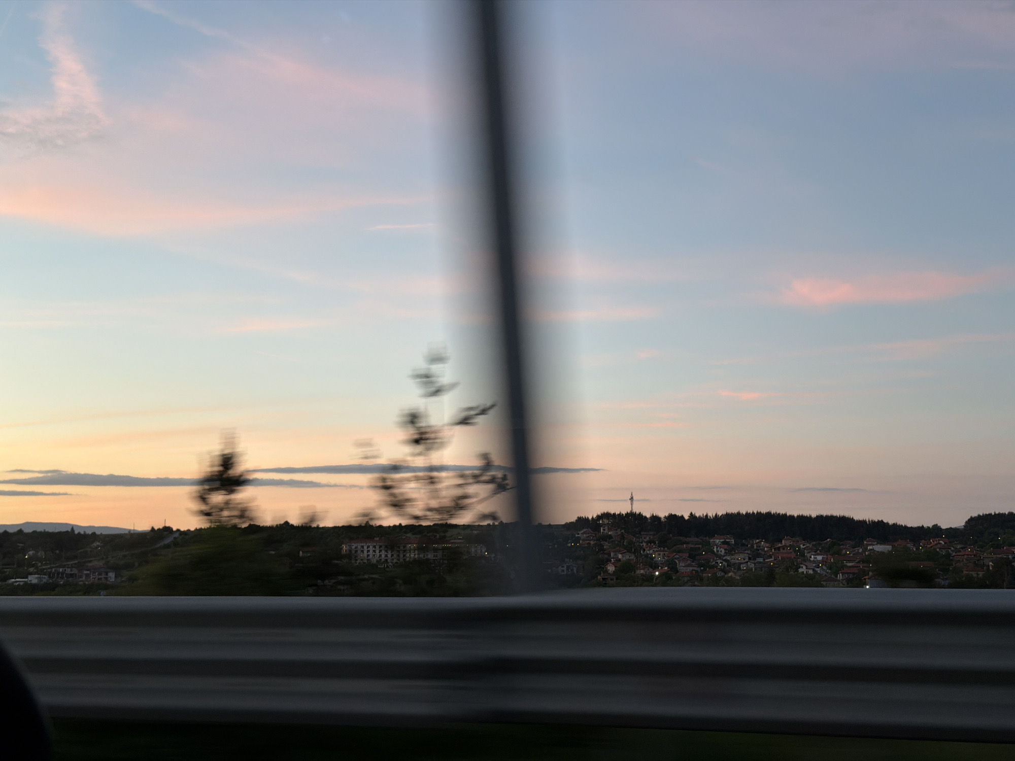 The image shows a view from a moving vehicle, likely a car, as evidenced by the blurred foreground object that appears to be a roadside barrier. Through the partially obscured view, a landscape unfolds with a clear sky tinted with shades of pink and orange, suggesting either sunrise or sunset.
In the midground, there is a settlement or town with houses clustered together, some of which are illuminated, implying it's either dusk or dawn. A densely forested area lines the horizon, adding depth to the scene. The blur from the vehicle's movement affects the sharpness of the vegetation and buildings visible between the obstruction in the foreground, but it's clear that the settlement is located on slightly elevated terrain. The image was taken in Baliovtsi, Bulgaria.