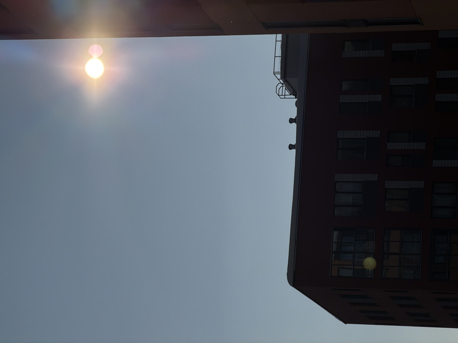 The scene depicts an urban landscape in Средний Постол, Russia, under a clear, light blue sky. The sun, a prominent bright white and yellow circular light source, is positioned in the upper right quadrant of the frame, emitting strong lens flare and a pinkish secondary flare artifact to its right.
In the lower left and central areas of the image, a multi-story building with dark reddish-brown exterior walls is visible, largely in shadow. This building features numerous rectangular windows arranged in a grid pattern; one window in the lower left reflects a yellowish circular lens flare artifact. The rooftop of this building displays two dark, circular ventilation structures.
To the right, a vertical section of a second, taller building with a lighter reddish-brown facade is partially visible. This building also appears to be in shadow and exhibits vertical architectural elements. Behind the roofline of the main reddish-brown building, a lighter colored structure with a metal railing or platform on its top is discernible, suggesting another building or rooftop installation further in the background. No individuals, overt actions, or interactions are present in the image.