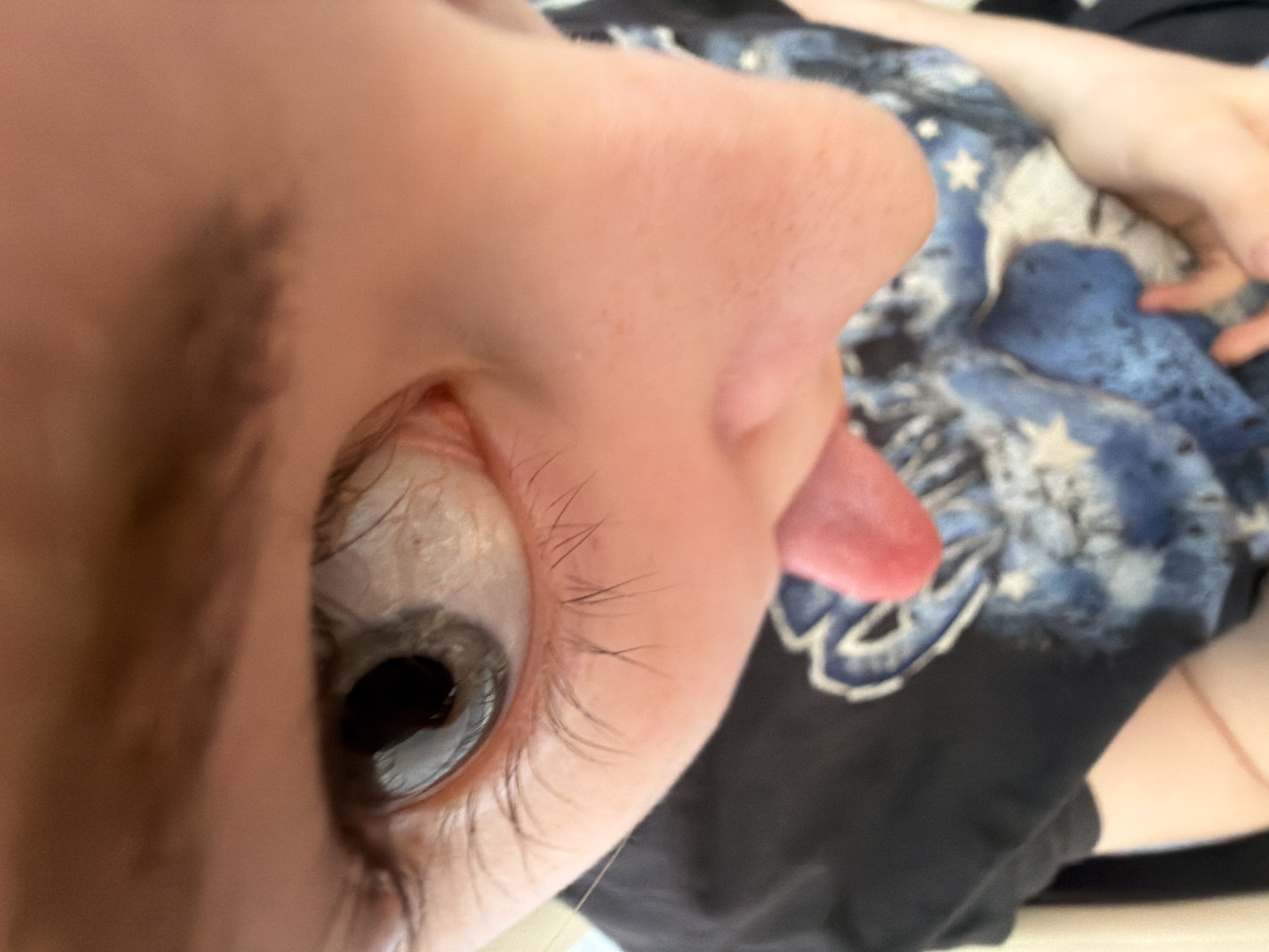 A close-up, disoriented image captures a person's face from an unusual angle, primarily focusing on their left eye, nose, and mouth. The person has light-toned skin. Their dark grey/blue eye is wide open, featuring visible eyelashes and fine red blood vessels in the white sclera, with a dilated pupil. Above the eye, a brow or forehead is partially visible, showing some fine facial hair. Part of the person's nose bridge and nostril are also in view. A small, pink tongue is extended slightly from the corner of the mouth, suggesting a playful or quirky expression.
In the blurred background, the person is wearing a black t-shirt adorned with a blue and white pattern featuring stars and cloud-like shapes, evoking a celestial or starry night theme. A portion of a hand or arm is also visible in the upper right. The lighting is soft and even, implying an indoor setting or diffused natural light. The overall perspective is intimate and abstract due to the extreme close-up and tilted angle. No visible text is present in the image. While the location context is Perm, Russia, no specific visual cues within the image confirm this setting.
