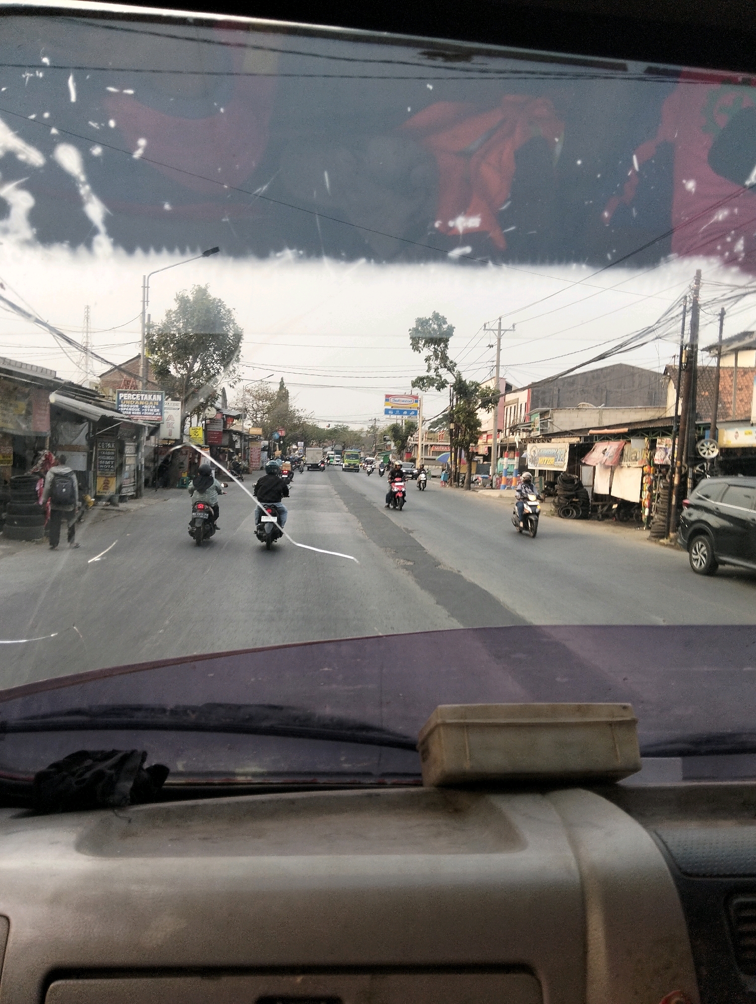 The image, taken from inside a vehicle looking out through the windshield, depicts a street scene in Semarang, Indonesia. The windshield is dirty with visible streaks and smudges, and a faint reflection of red fabric or objects is present. The foreground shows the interior dashboard of the vehicle, with a dark cloth on the left and a small, tan, rectangular box placed on the dashboard on the right.
The street is paved and appears to be a moderately busy road with several motorbikes in motion. On the left side of the road, there are buildings that look like shops or small businesses, with awnings and signage. One sign is partially visible and appears to be in Indonesian, with text such as "PERCETAKAN" and "CETAK OFFSET". There are stacks of tires visible outside one of these establishments. A person with a backpack is walking on the sidewalk.
On the right side of the road, more buildings are visible, including what appears to be a store with a blue and white sign that resembles "Indomaret". Another building has signage that is difficult to read. A black SUV is parked on the side of the road.
Several streetlights and utility poles with numerous wires are present along the street, indicating urban infrastructure. The sky is overcast, suggesting either early morning, late afternoon, or a cloudy day. The overall environment is a typical street scene in a developing urban area, with a mix of commercial establishments and residential buildings, bustling with traffic, primarily motorbikes.