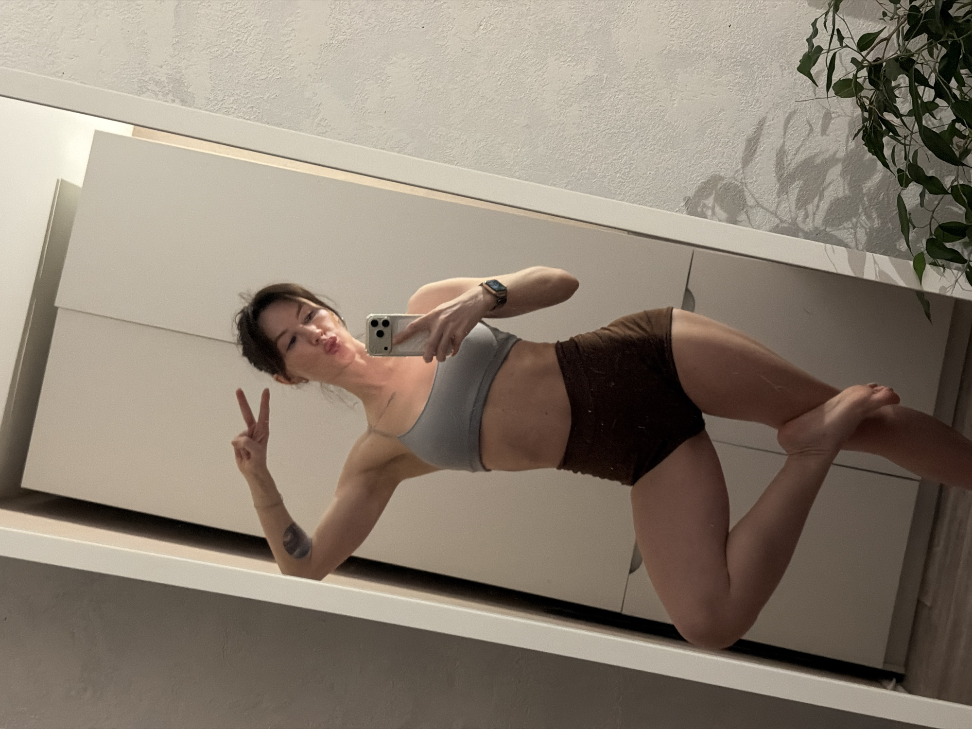 A woman takes a selfie in a mirror, leaning against a shelf or counter with white cabinets behind her. She is wearing a grey sports bra and brown shorts. She is holding a phone in her right hand, displaying a smartwatch on her wrist, and making a peace sign with her left hand. A tattoo is visible on her left forearm. A plant is situated near the top right of the frame. The location or the city of Perm, Russia cannot be identified from this picture alone.