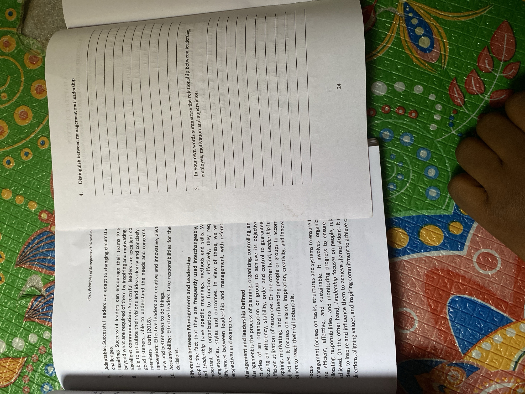 An open textbook, displaying academic content on "Basic Principles of Entrepreneurship" and detailing "Management and Leadership" concepts including leadership qualities and definitions, rests on a surface. The visible page, numbered 24, includes questions with blank lines for responses. The textbook is positioned on a colorful, embossed plastic mat featuring a repeated geometric and organic pattern in shades of green, blue, yellow, red, and white. A light-skinned human hand, with fingers slightly curled, is partially visible in the lower-left foreground, resting on the patterned mat. The scene suggests an indoor study or reading environment in Nsukka, Nigeria.