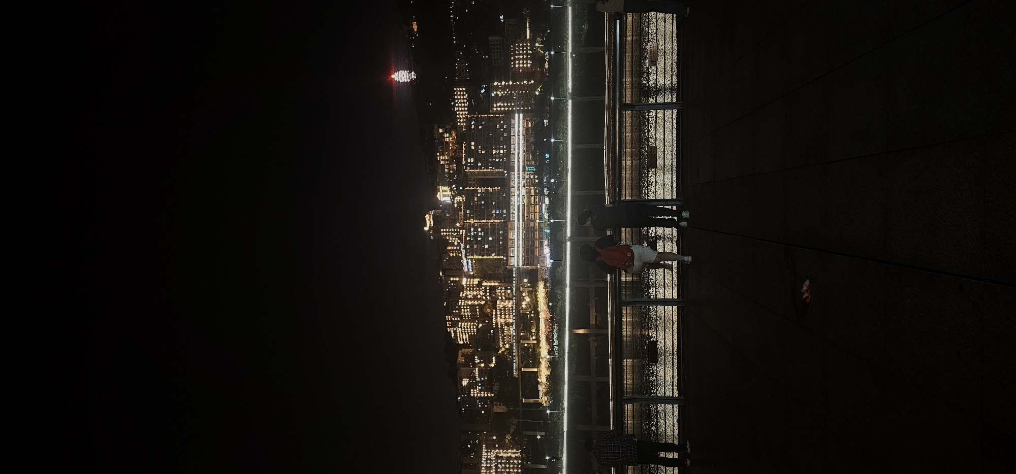 From an elevated position, a view of 江北城街道, China at night is depicted. A group of people stand on a platform, observing the cityscape. The city is lit with numerous lights, creating a vibrant glow against the dark sky. A body of water reflects some of the city lights. In the distance, there's a lit boat. The atmosphere is dark and night-time, illuminated primarily by the lights of the city and the reflection on the water.