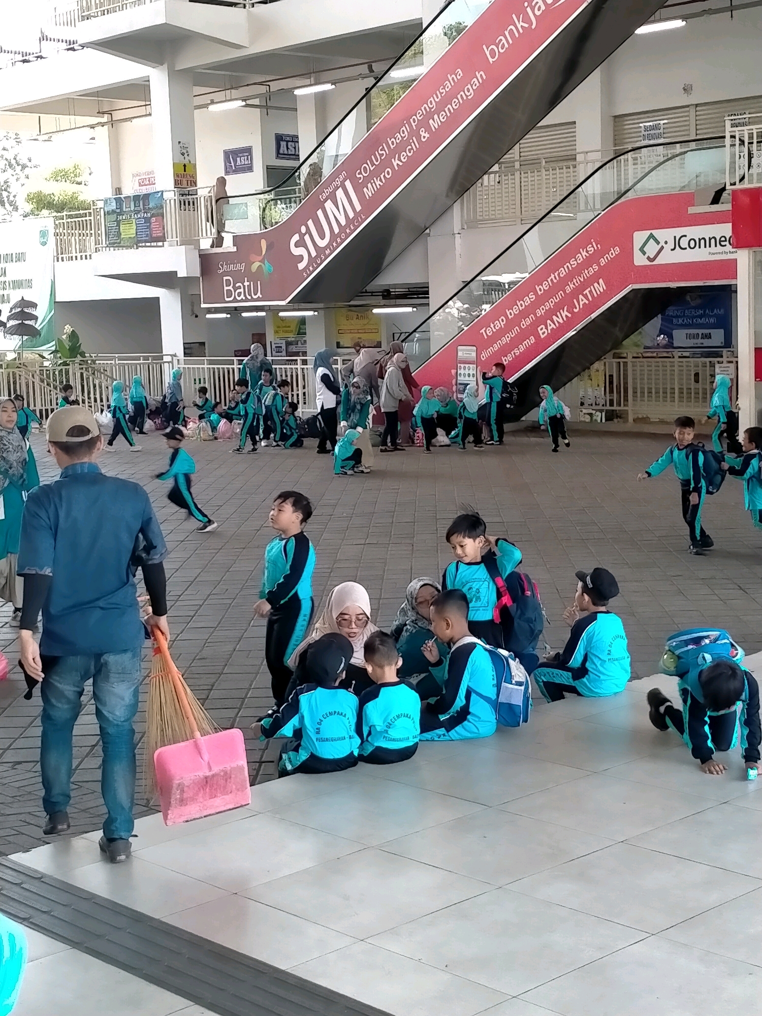 The image captures a scene within a public building, likely a transportation hub or market, in Batu, Indonesia. The architecture suggests an indoor-outdoor space with a covered walkway leading to escalators. A large red advertisement for "SiUMI" (Siklus Mikro Kecil) and "BANK JATIM" dominates the background, promoting financial services.
In the foreground, a man in a blue shirt and jeans is holding a broom and dustpan, appearing to be cleaning the tiled floor. A group of children, mostly wearing matching teal and black uniforms, are scattered around. Some are sitting on the floor, others are standing, and a few are in motion. The children seem to be engaged in various activities, with some interacting with each other, while others appear to be resting or waiting.
The environment includes visible signage, railings, and potted plants, contributing to a sense of a busy public space. The presence of children in uniform suggests a school outing or an organized event. The overall atmosphere appears to be one of casual activity and movement within a public facility.