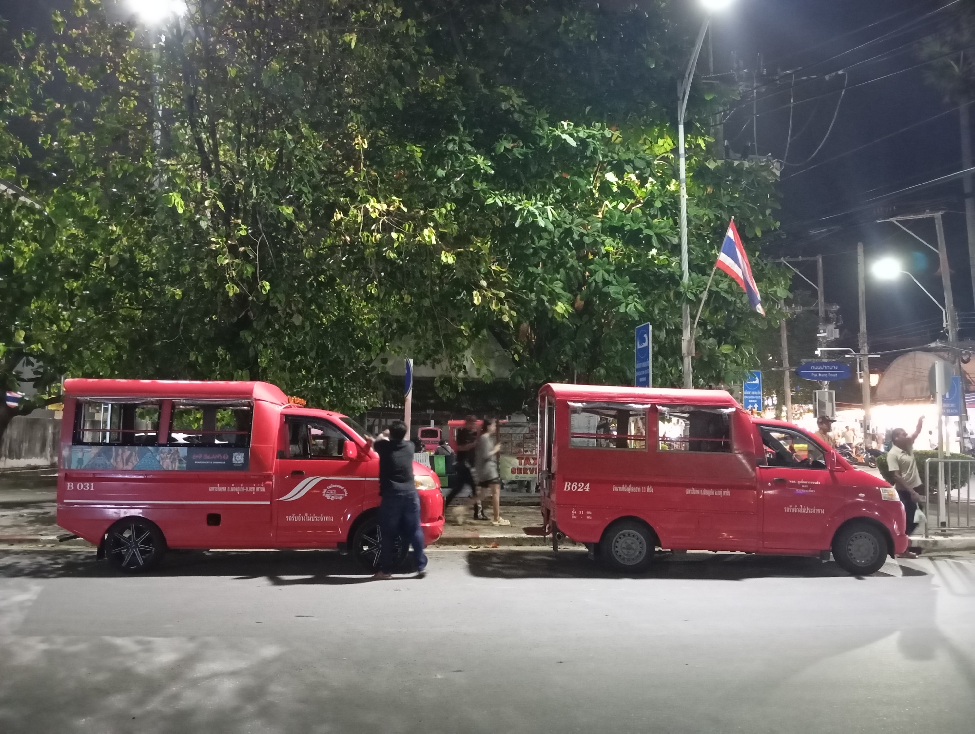 Two bright red songthaews, a type of shared taxi common in Thailand, are parked on a street in Karon, Thailand. The songthaews have covered seating areas in the rear, characteristic of this vehicle type. The one on the left has the number "B 031" on its side, and the one on the right displays "B624". A Thai flag is visible in the background, attached to a pole, fluttering slightly. The scene is illuminated by streetlights, casting shadows and highlighting the foliage of a large tree that dominates the upper portion of the image. There are a few people visible, some appearing to interact near the songthaews, with motion blur suggesting they are in motion. The overall setting suggests it is nighttime, with the ambient light illuminating the street and surrounding area.