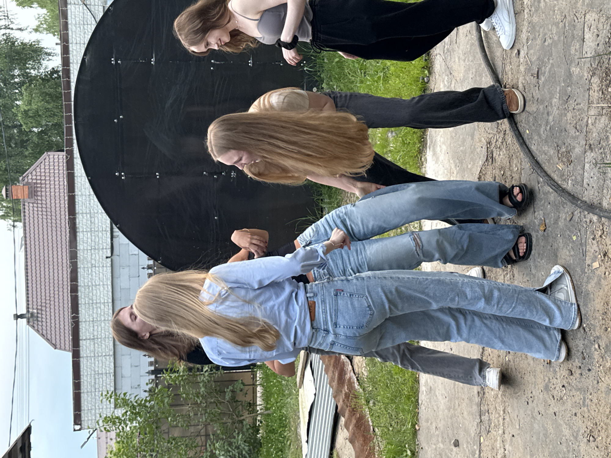 The image captures a group of young women outdoors, gathered around a large, dark, circular object that appears to be a satellite dish or a similar structure. The scene is set in a somewhat rustic environment with visible grass, concrete ground, and glimpses of residential buildings with tiled roofs. The sky suggests it's daytime, possibly overcast or late afternoon given the diffused light.
The women are dressed casually in jeans and tops, suggesting a relaxed atmosphere. Their poses and interactions imply they are engaged in some form of activity, possibly a game or simply socializing. One woman in the foreground, wearing a light blue shirt and jeans, is looking down and to her left, her hair falling forward. To her left and slightly behind her, another woman with long blonde hair is also looking downwards. Further back, a woman with brown hair in a grey tank top and black pants is looking to her right, and another individual in a light-colored shirt and dark pants is seen with their back to the camera.
Notable details include the worn appearance of the jeans worn by some of the women, a black scrunchie on one woman's wrist, and a visible red tag on the back pocket of the woman in the light blue shirt, possibly indicating a brand. The ground is a mixture of cracked concrete and dirt, with some patches of grass. The overall impression is one of youthful energy and camaraderie in an informal outdoor setting. The location context of Perm, Russia, adds a layer of geographical understanding to the scene. There is no discernible text in the image.