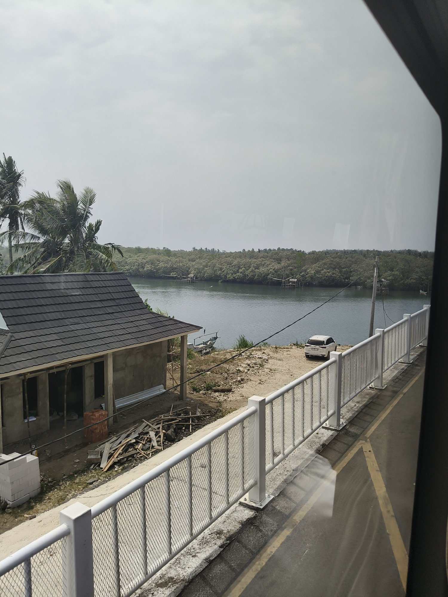 A daytime, wide-angle view captures a coastal scene in Desa Cijulang, Indonesia. In the foreground, a grey, unfinished concrete building with a dark grey tiled roof is under construction, with piles of bricks and wooden planks scattered nearby. A white metal fence with a diamond mesh pattern runs along the right side of the frame, separating the construction area from a paved pathway marked with yellow lines. Beyond the fence, a calm, dark body of water is visible, reflecting the overcast sky. On the opposite bank of the water, a dense forest of green trees lines the shore, with several small wooden structures on stilts rising from the water. A white SUV is parked on a sandy patch of the opposite shore. Two palm trees with green fronds are visible in the upper left corner of the image, partially obscuring the cloudy sky. The scene suggests a developing coastal area with both construction and natural elements.