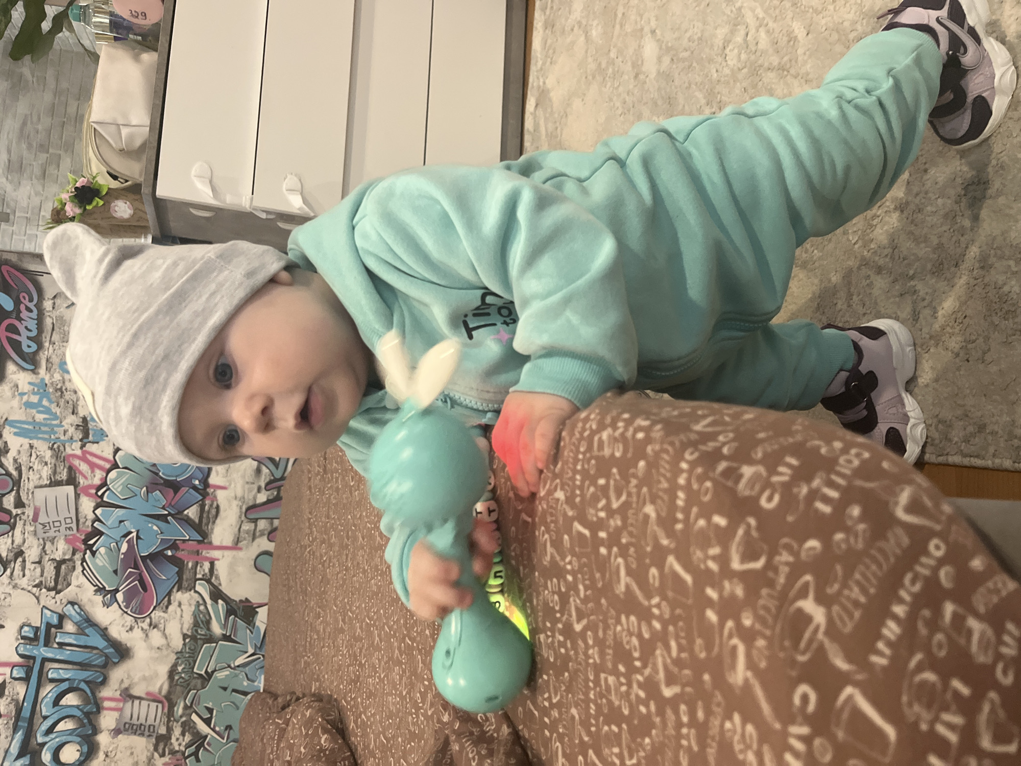 A baby wearing a gray hat and a light blue hooded tracksuit is positioned on a patterned brown surface. The baby is holding a blue toy in its hands. The background includes a patterned wall and a white cabinet. The location is not discernible from the image.