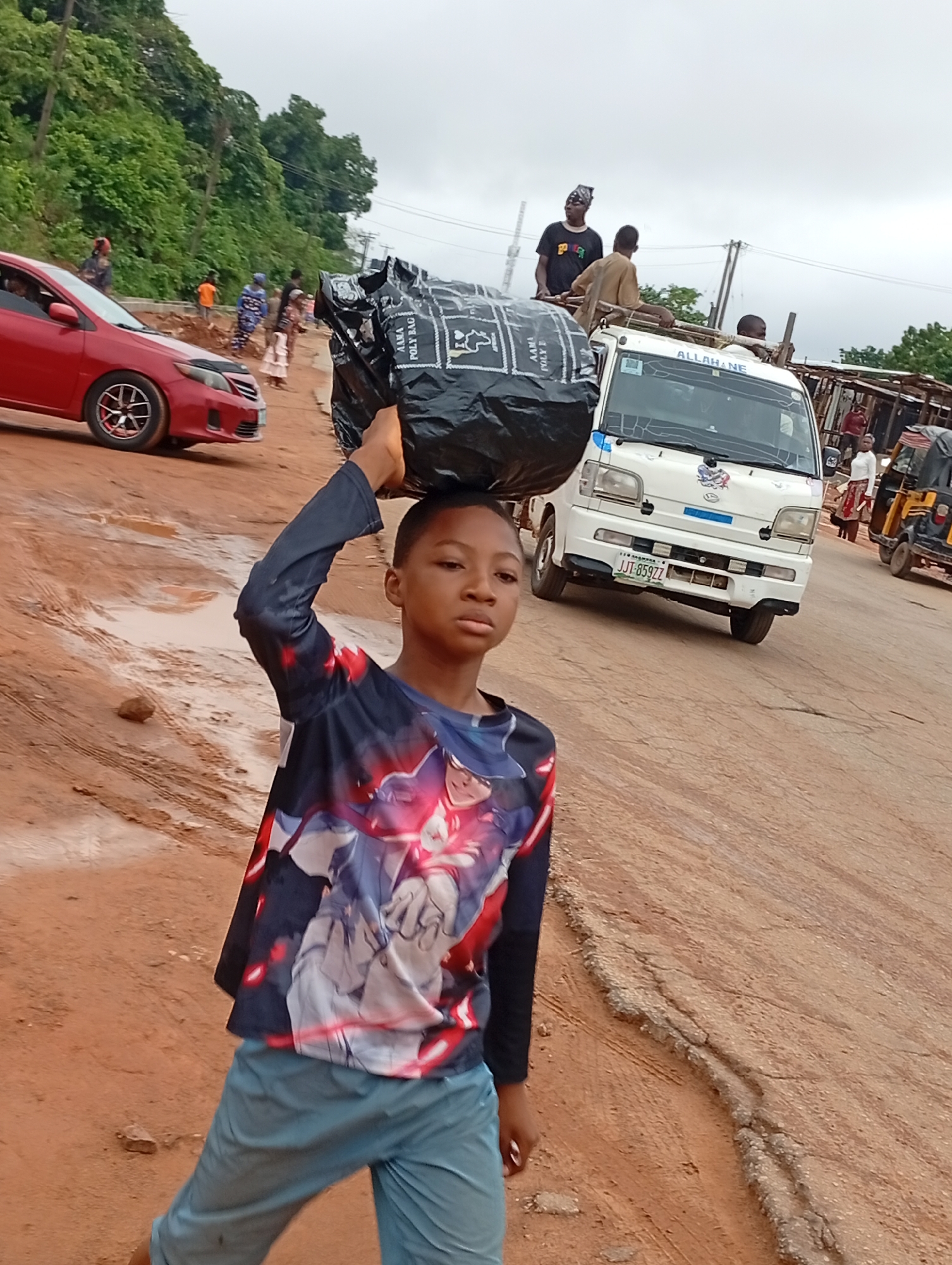 In Nkwelle Ezunaka, Nigeria, a young boy with a serious expression carries a large black bag on his head, steadying it with his hand. He wears a long-sleeved shirt with a graphic design and light blue pants. The road is unpaved, reddish-brown, and shows signs of disrepair. Behind the boy, there is a red car parked on the left and a white van with people standing on its roof and in the back on the right. Other people are walking along the road, and there's a three-wheeled vehicle to the right. Green trees and vegetation are visible in the background.