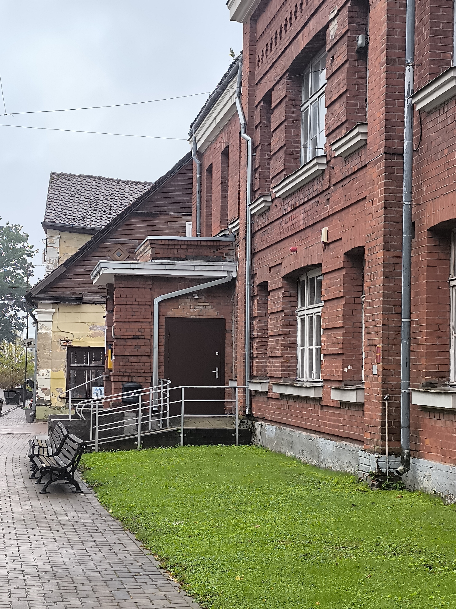 The image depicts a street scene in Kuldīga, Latvia, on an overcast day. In the foreground, a paved walkway made of reddish-brown brick leads towards the viewer. Two dark, wooden park benches with metal legs are situated on the left side of the walkway, facing away from the viewer. Adjacent to the walkway on the right is a patch of vibrant green grass.
Behind the grass and walkway, a large, red brick building with white trim and arched windows dominates the right side of the frame. This building appears to be an older structure, with visible details such as downspouts, architectural embellishments between the windows, and a concrete foundation.
Further back and to the left, a slightly older, lighter-colored building with a weathered facade and a dark, tiled roof is visible. This building has a peaked roofline and appears to be attached to or adjacent to the red brick structure. A brown wooden door with a metal handle is visible on the red brick building, accessible via a ramp with metal railings. A small poster with a drawing of a face is affixed to the railing.
The overall environment suggests an urban setting, likely a street or public area in Kuldīga. The sky is a uniform grey, indicating a cloudy day. No people are visible in the immediate scene.