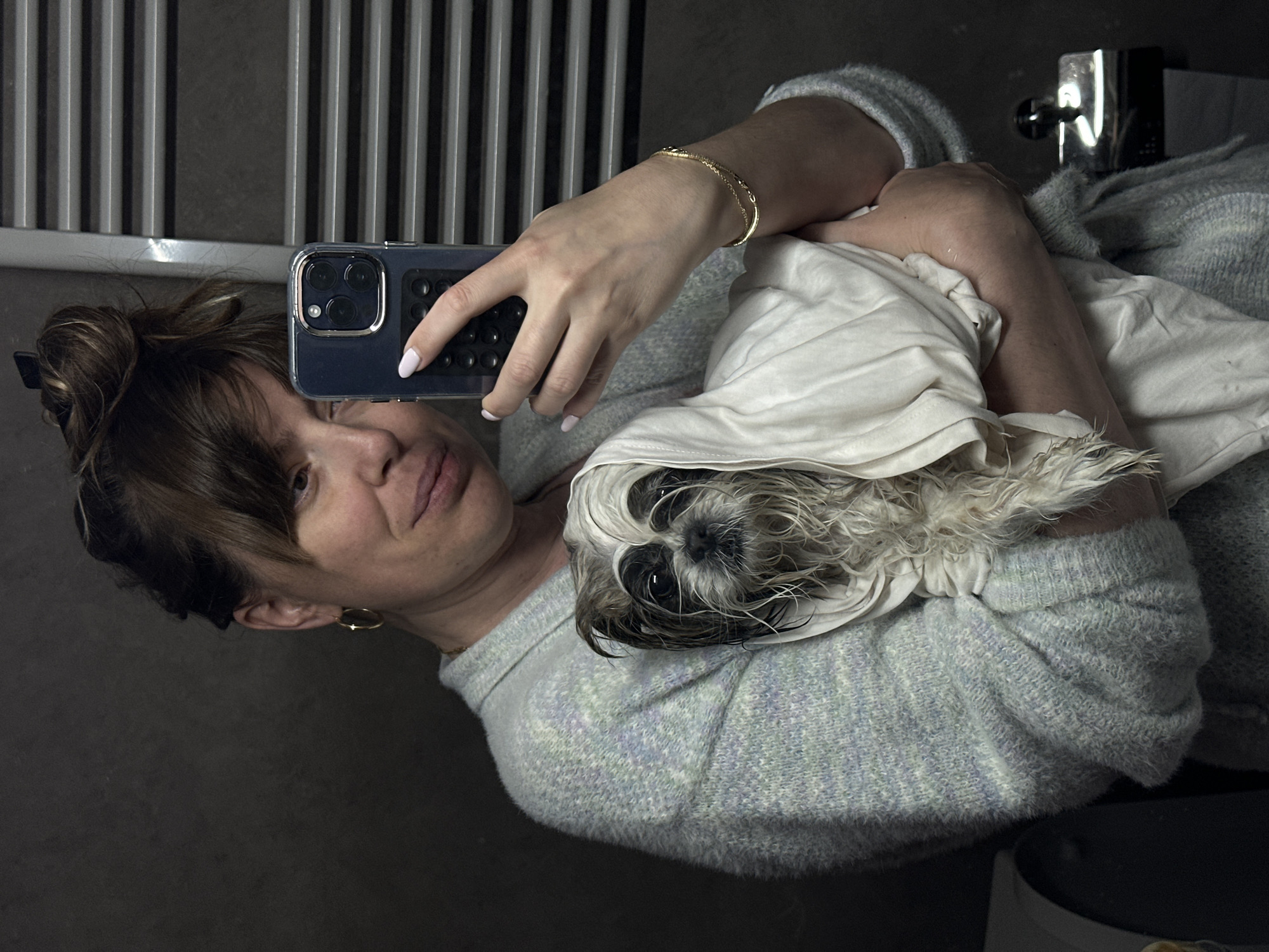 A woman is taking a selfie with her dog in a bathroom. She is holding a smartphone with her right hand. The dog is wrapped in a white towel and rests in her left arm. The woman is wearing a light grey sweater and gold jewelry (bracelet, earrings). Her hair is in a messy bun. In the background is a bathroom radiator with vertical bars and a sink faucet. The location is inferred to be Tbilisi, Georgia.
