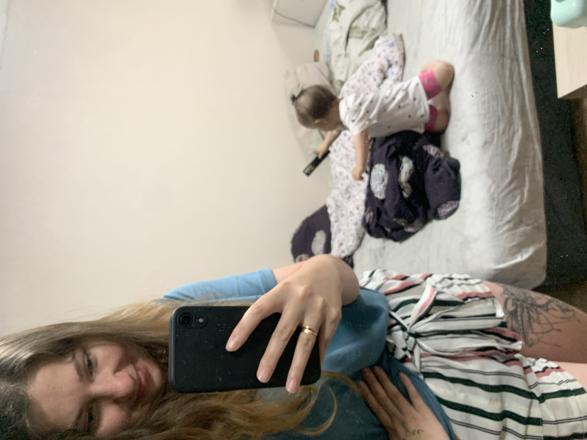 The image is a mirror selfie taken indoors, likely in a bedroom. In the foreground, a woman is visible from the chest down, holding a black smartphone to take the picture. She is wearing a teal t-shirt and striped shorts. Her left hand rests on her stomach, and her right hand is holding the phone, displaying a gold ring. On the bed in the background, a toddler is seated, holding a black remote control and looking away from the camera. The bed is unmade and appears to be covered with various blankets and pillows. The overall environment suggests a casual, domestic setting. The image does not provide any information that would allow for its specific location in Chaikovsky, Russia.