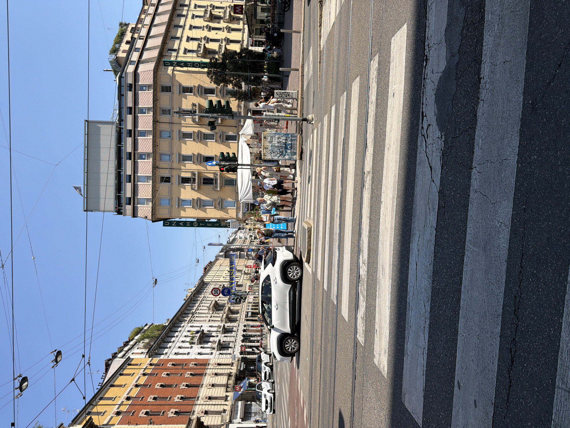 The image captures a street scene in Milan, Italy, likely near a busy intersection in an urban area. The foreground features a wide pedestrian crossing with prominent white stripes on asphalt. Beyond the crossing, a white SUV is stopped, positioned to allow pedestrians to cross. Several people are visible on the sidewalk and near the crossing, some appearing to be waiting to cross the street, while others are engaged in conversation.
The background is dominated by multi-story buildings that exhibit classic Milanese architecture, characterized by their yellow and brick facades, numerous windows, and balconies. One building prominently displays a sign for "UNAHOTELS". Overhead, a complex network of electrical wires for trams or trolleybuses crisscrosses the sky, a common sight in Milan. The sky is clear and blue, indicating a sunny day. The overall impression is of a vibrant city street with active pedestrian and vehicular movement.