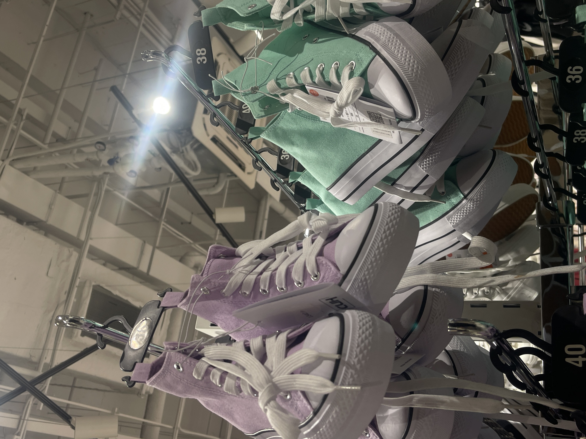 The image shows a display rack of high-top canvas sneakers in a retail setting. The shoes are various colors, including mint green and lavender, and are hanging by hangers. The hangers also display the sizes. The background shows a commercial or retail space with a high ceiling and exposed piping. The location is Kommunarka, Russia.